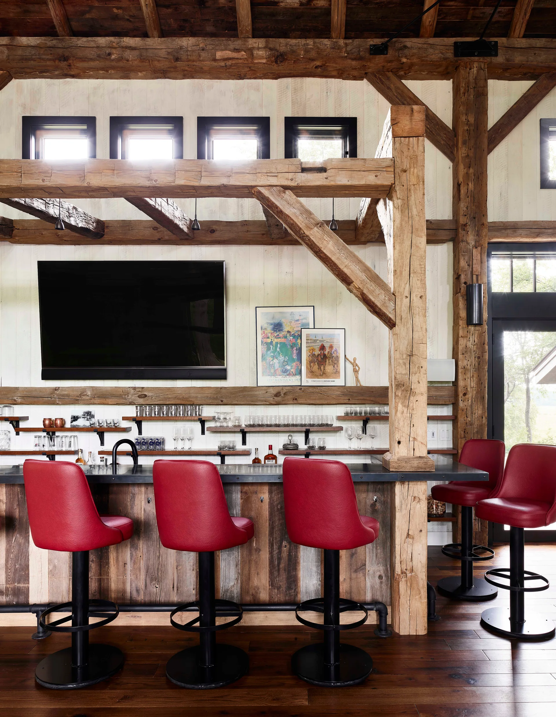 A rustic home bar featuring a metal-topped wood counter, red bar stools, open glass shelves, and exposed timber frame beams.