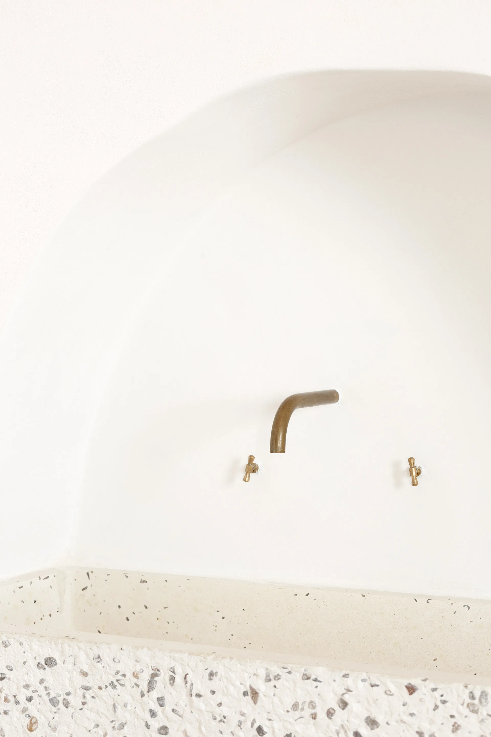A wall-mounted brass faucet and two cross handles are installed above a textured terrazzo sink basin in a white arched plaster wall alcove.