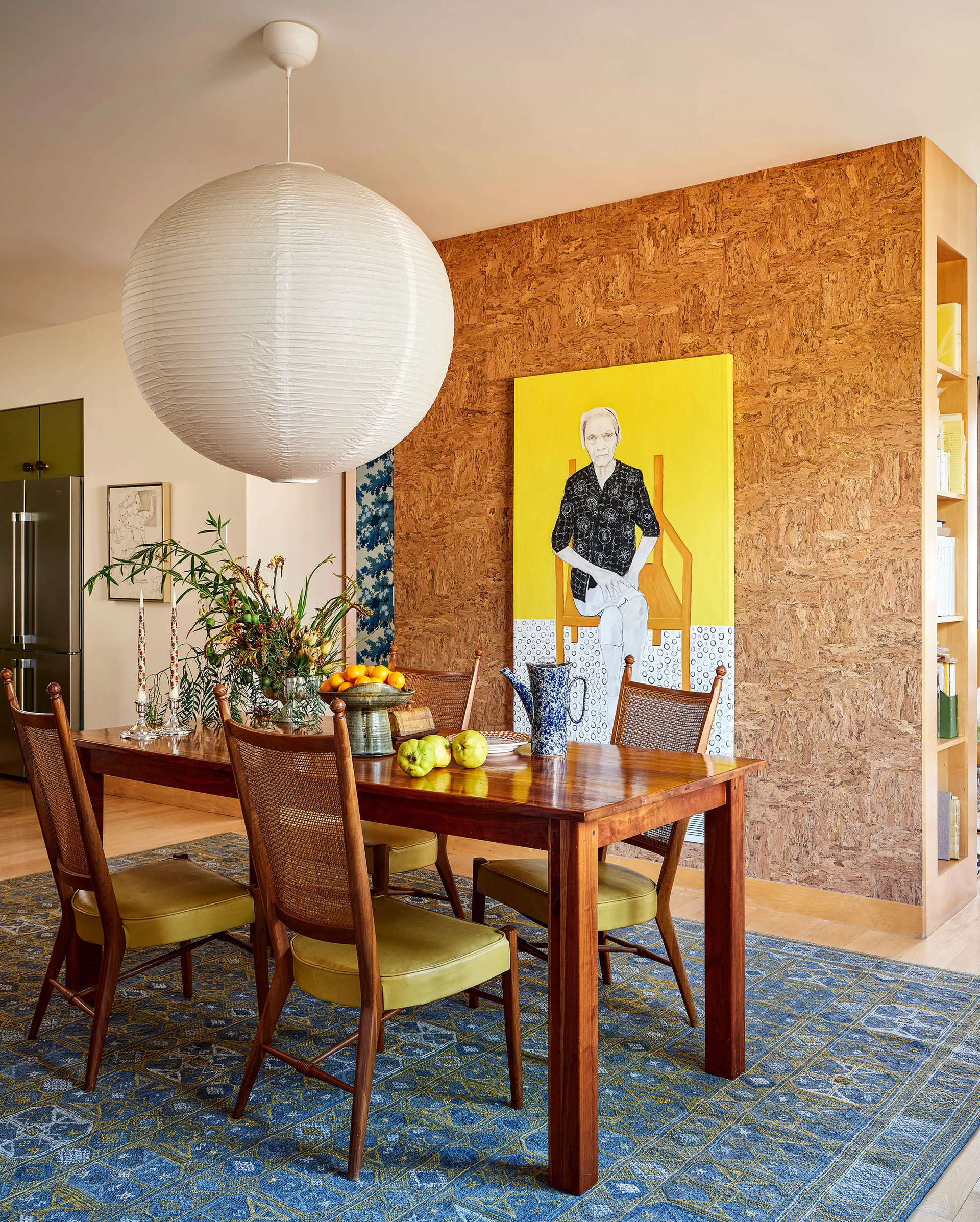 A wooden dining set with cane-back chairs rests on a patterned blue rug in front of a textured cork wall decorated with a large yellow painting.