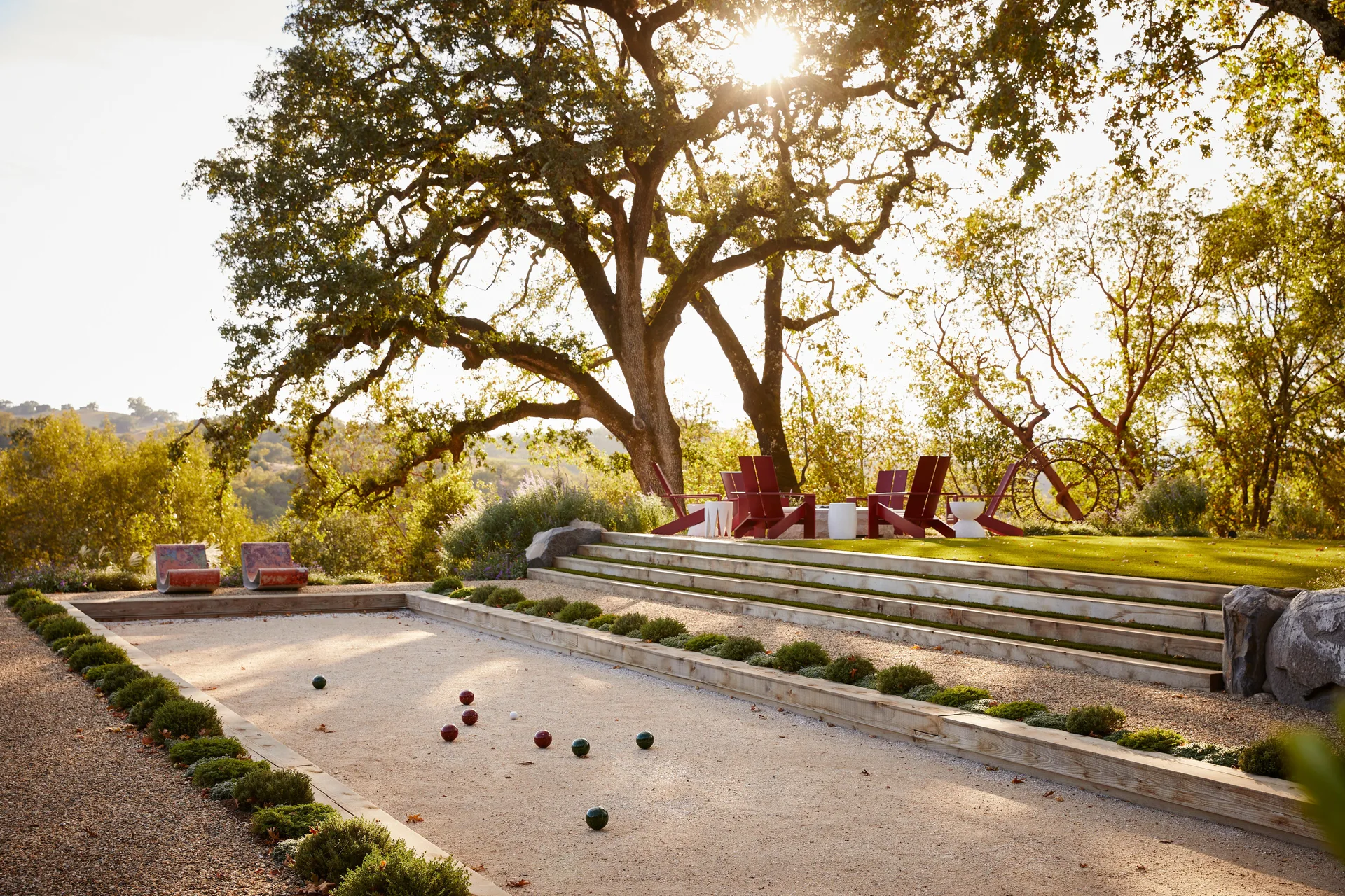 A backyard bocce ball court next to tiered wooden steps leading to a grassy lawn with red Adirondack chairs under a large oak tree.