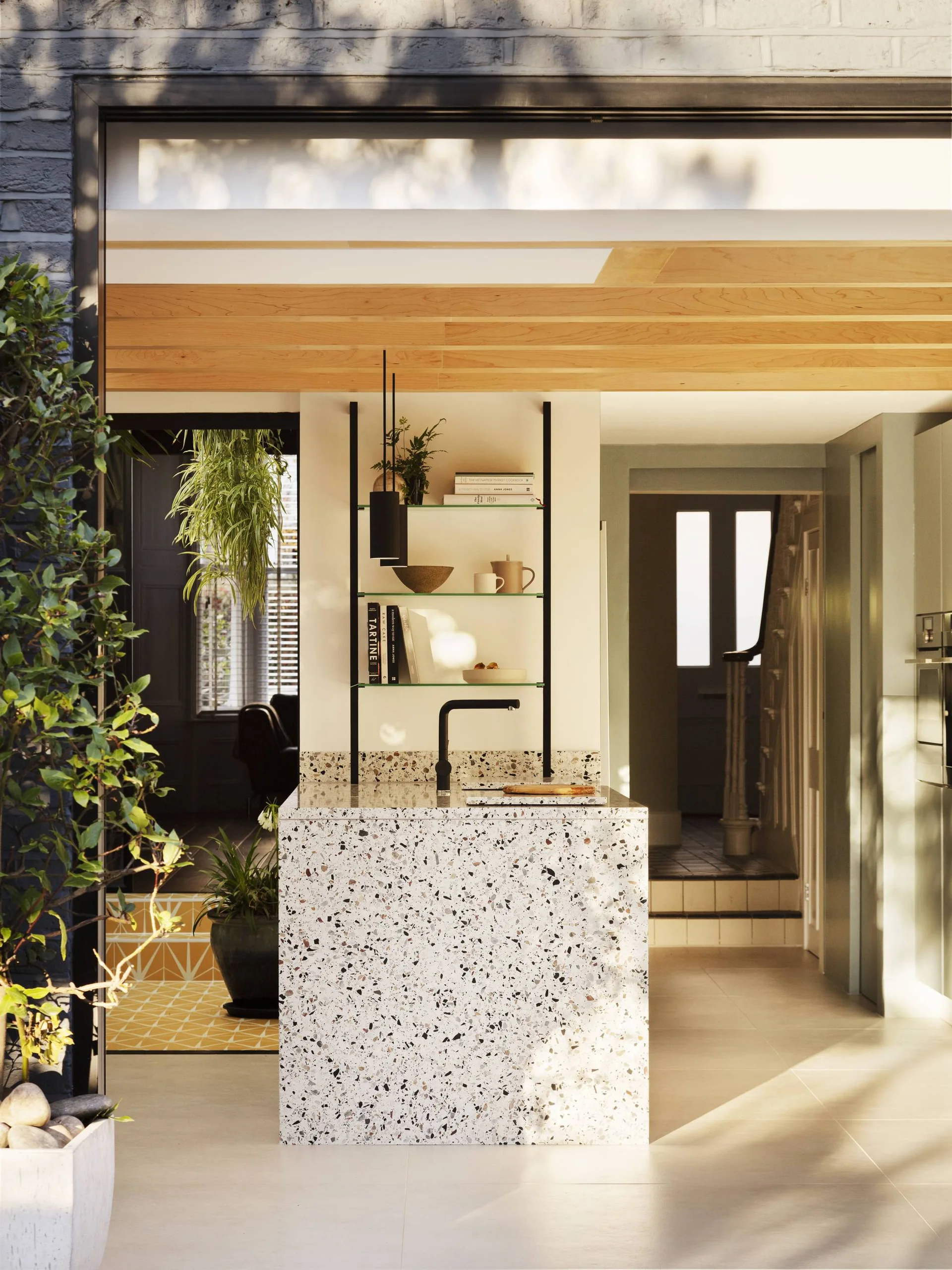 A sunlit kitchen features a speckled terrazzo island with a black faucet, open glass shelving, and a natural light wood-paneled ceiling.