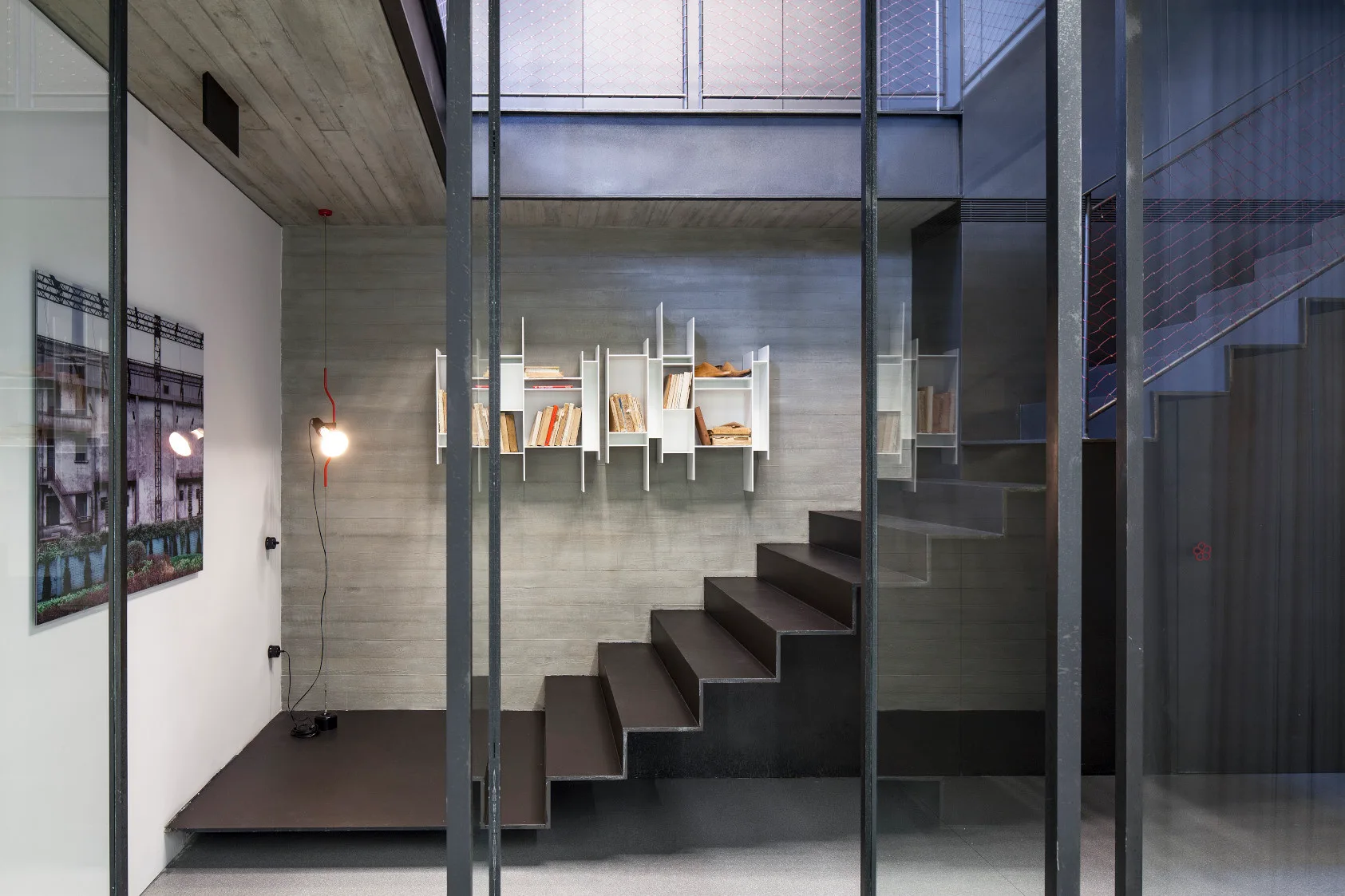 A black metal staircase stands against a grey board-formed concrete wall featuring a white modular shelf with books behind vertical glass partitions.