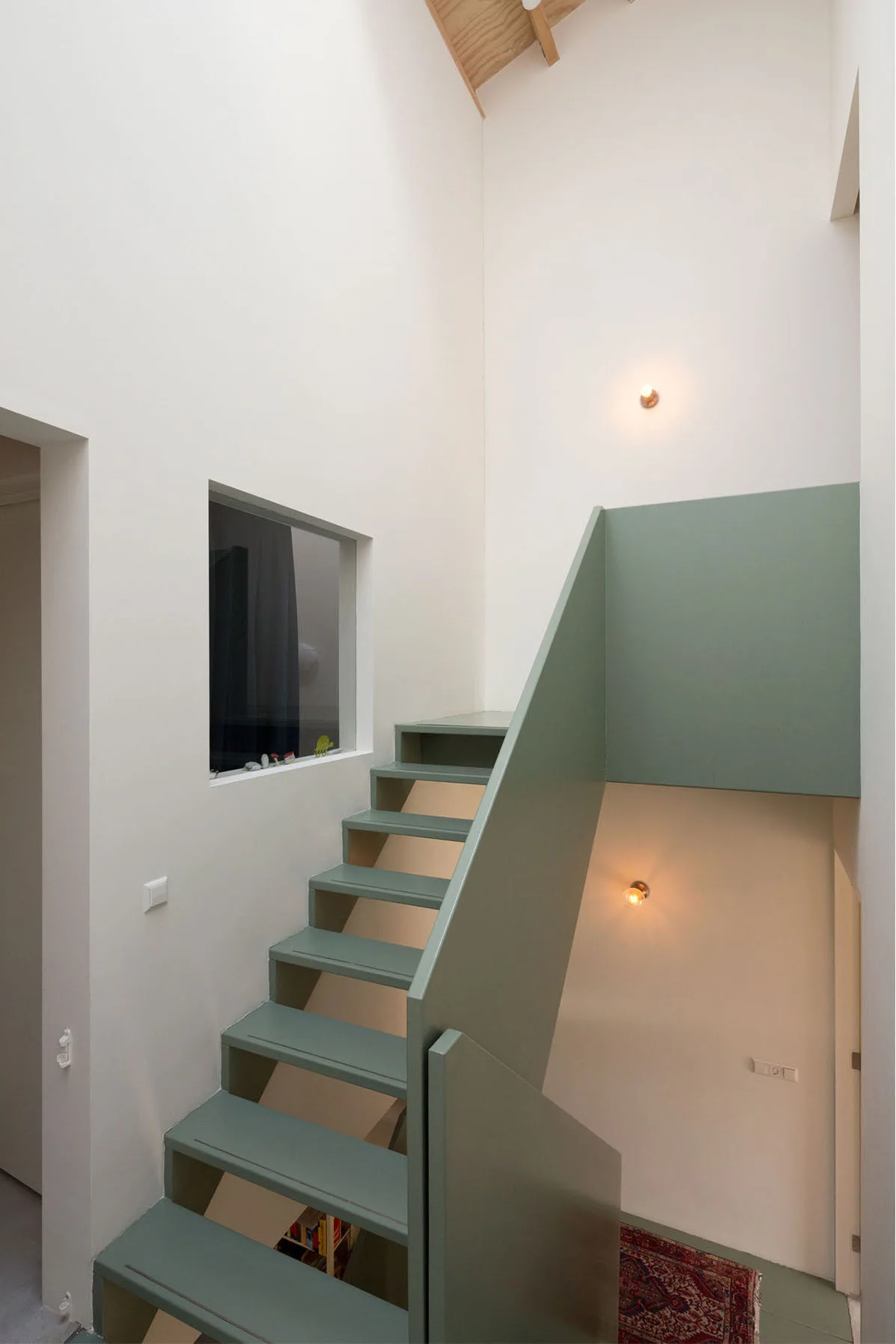 A minimalist sage green staircase with solid panel railings rises against off-white walls illuminated by warm circular wall sconces.