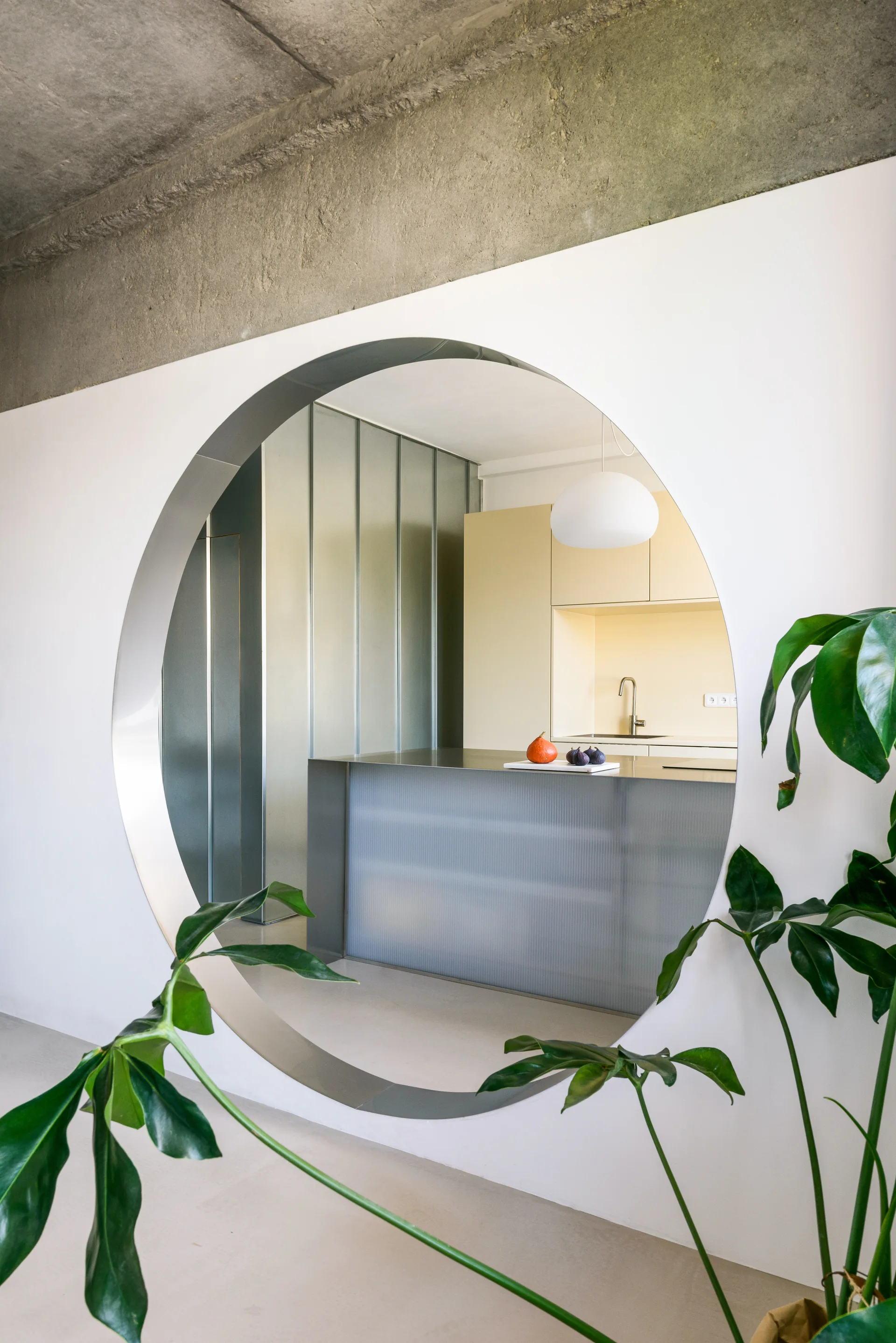A view through a large circular cutout in a white wall reveals a minimalist kitchen with pale yellow cabinets, a metal island, and a raw concrete ceiling.