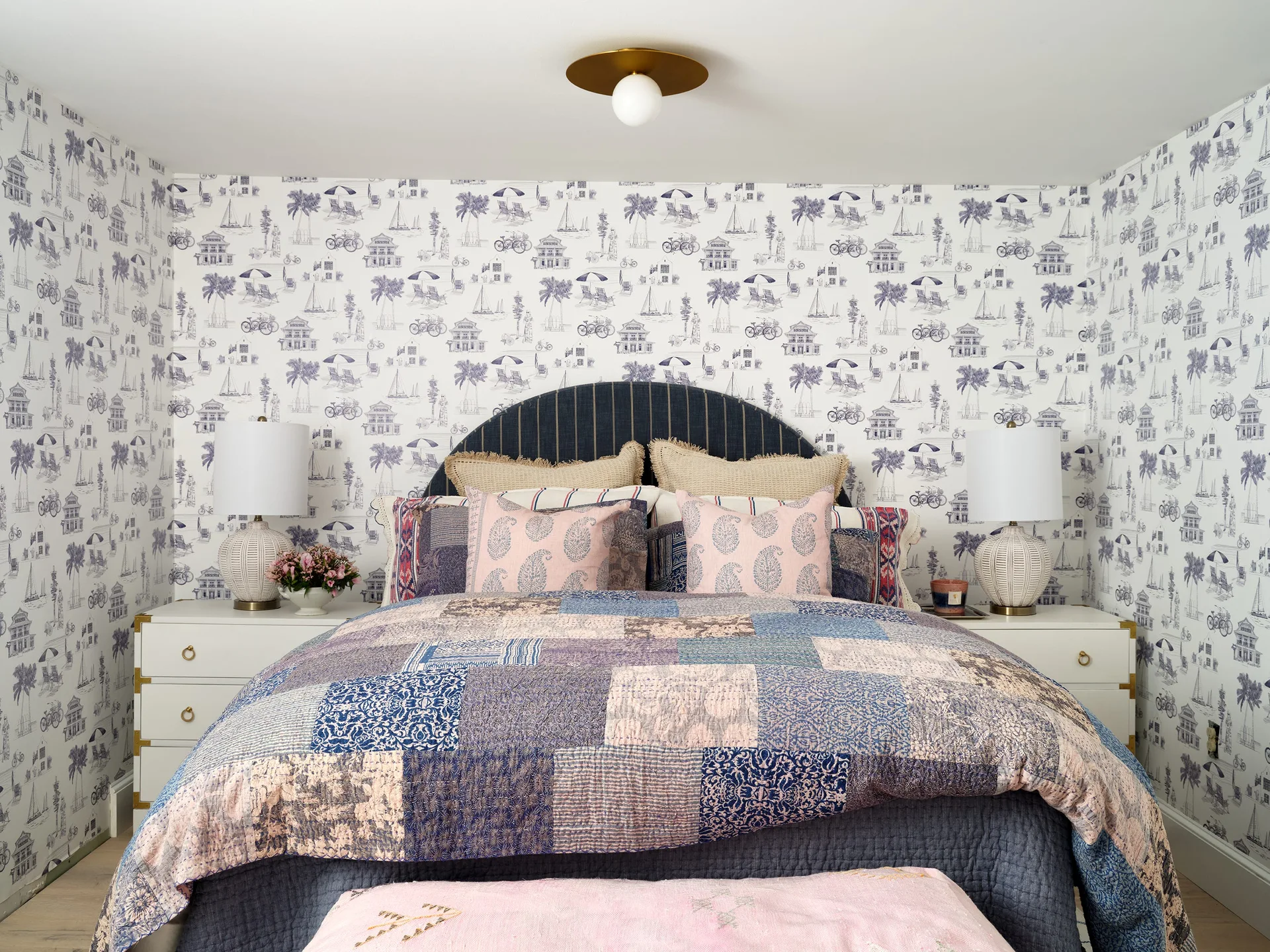 A symmetrical bedroom features illustrated blue-and-white wallpaper, a dark arched headboard, and matching white nightstands with textured table lamps.