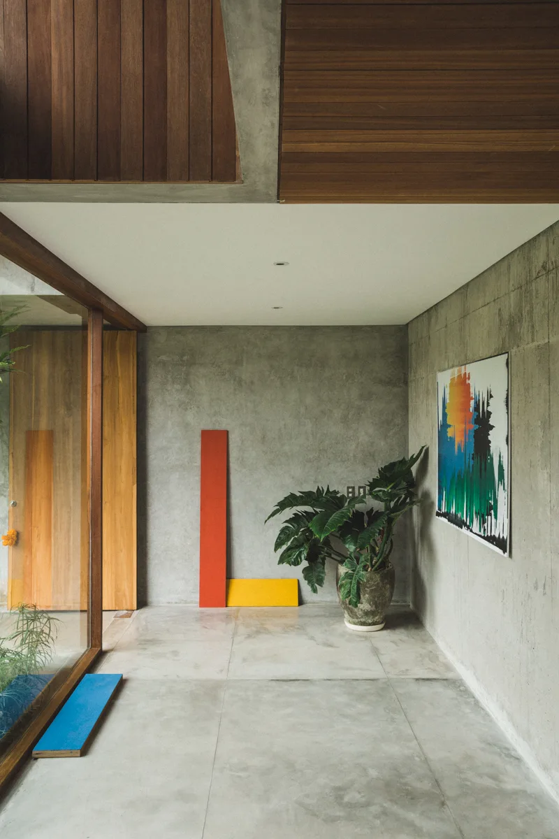 A minimalist concrete entryway features a wood-slatted ceiling, a large potted plant, and leaning red and yellow art panels on the floor.