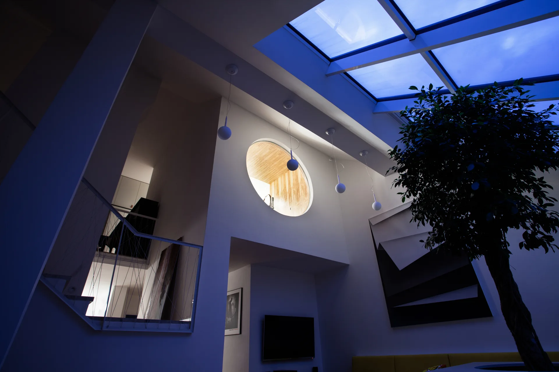 A double-height modern room features a large overhead grid skylight, white globe pendant lights, and a circular interior window lined with warm wood.