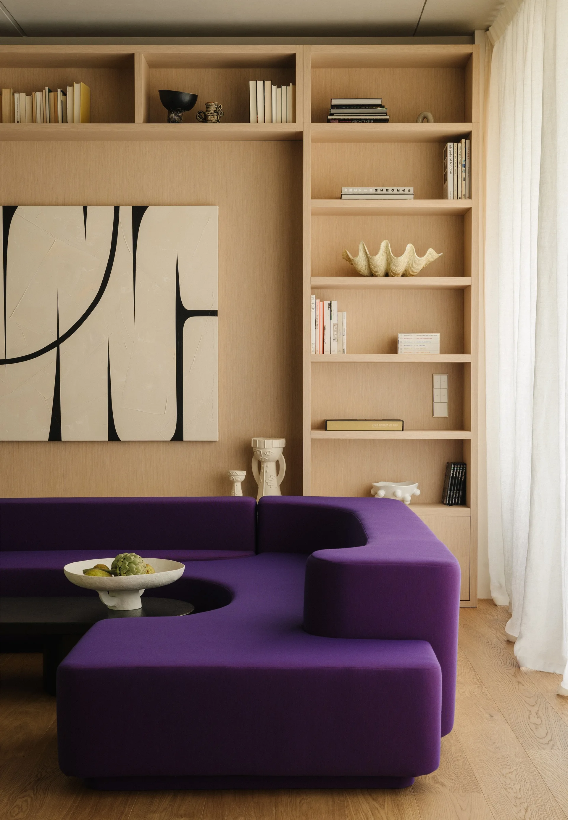 A modern living room featuring a curved purple modular sofa set against a light oak wood paneled wall with built-in shelves and abstract art.