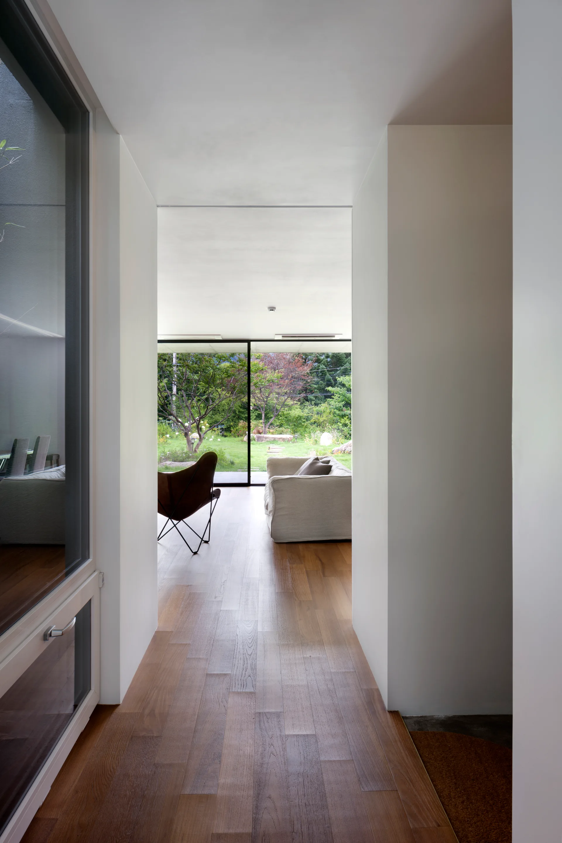A hallway with wood flooring leads to a bright living room with a sofa, leather butterfly chair, and a sliding glass door facing a garden.