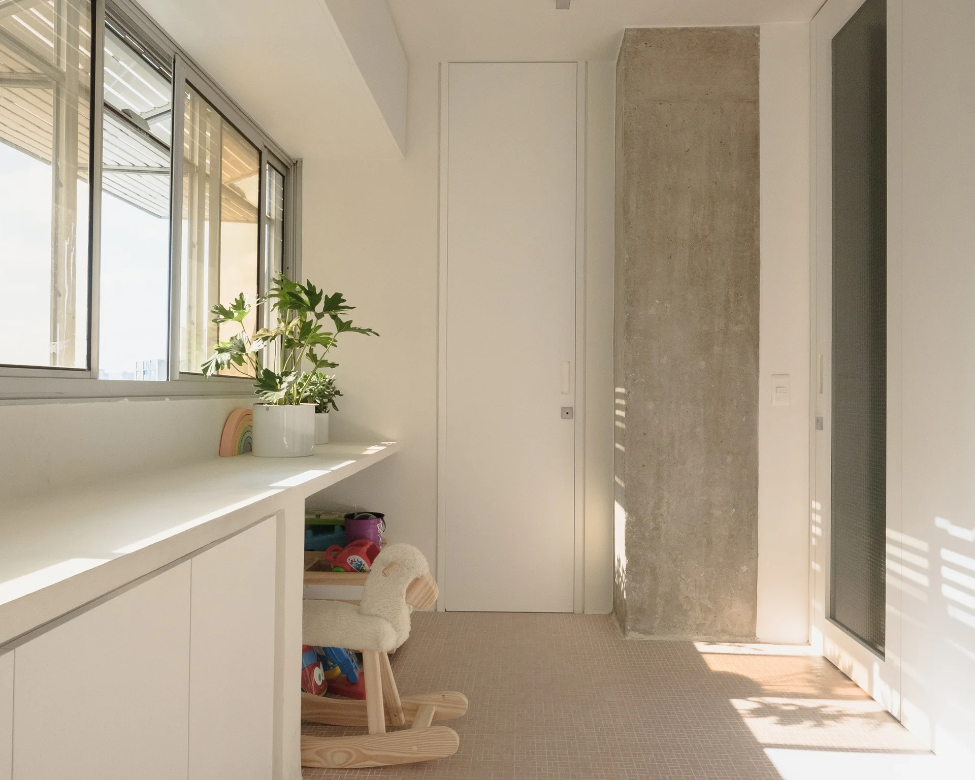 A bright room features a long white built-in desk under a window, a child's rocking horse, and an exposed raw concrete column.