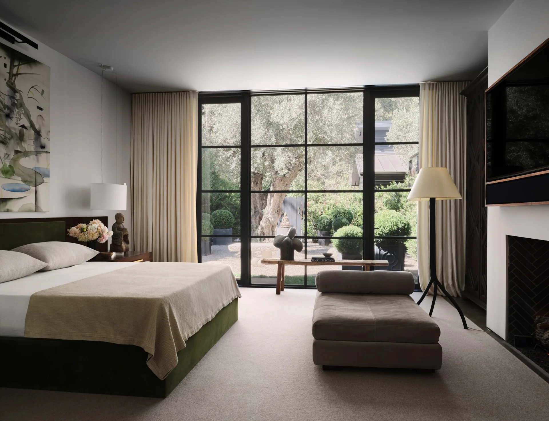 A modern bedroom features a green upholstered bed, a grey chaise lounge, and a large black metal grid window looking out to a garden.