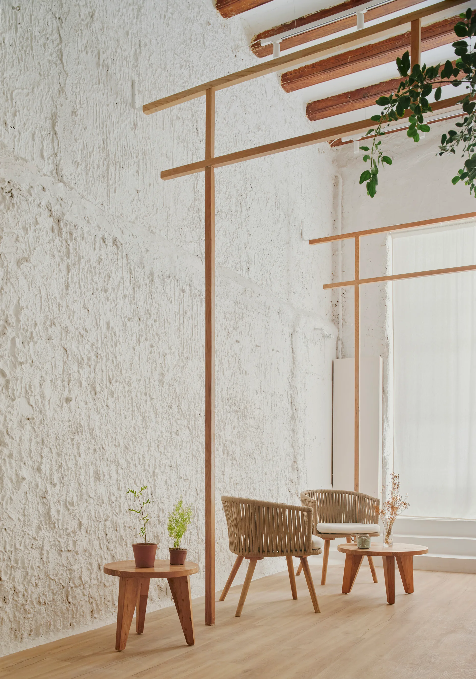 Two woven armchairs and round wooden side tables are arranged in a minimalist room with a highly textured white plaster wall and slim timber framing.