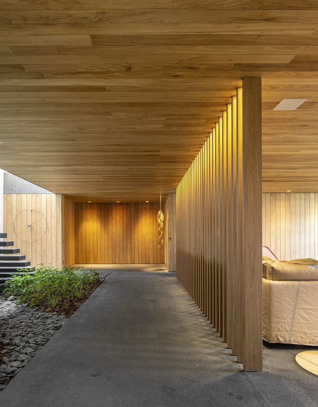 A modern entryway features a slatted wood partition wall, a wood-paneled ceiling, dark stone flooring, and an indoor garden bed with green plants.