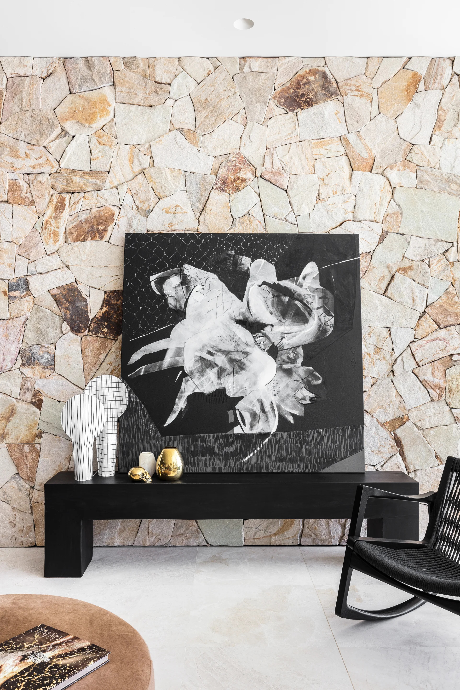 A minimalist black console table stands in front of a textured stone wall, decorated with a large abstract painting and two grid-patterned lamps.