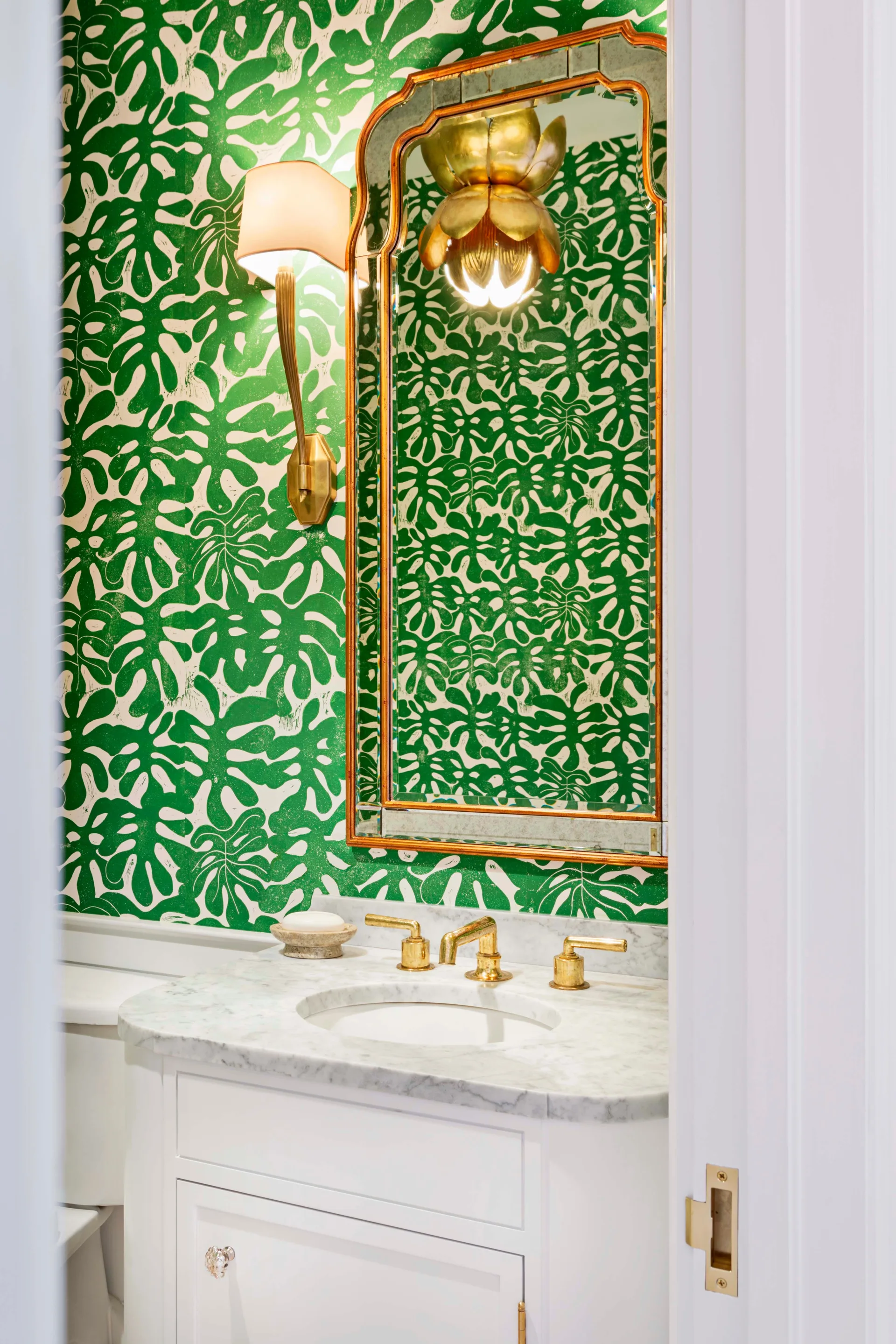 A small powder room featuring bold green and white leaf-patterned wallpaper, a white marble vanity with brass faucets, and a gold framed mirror.