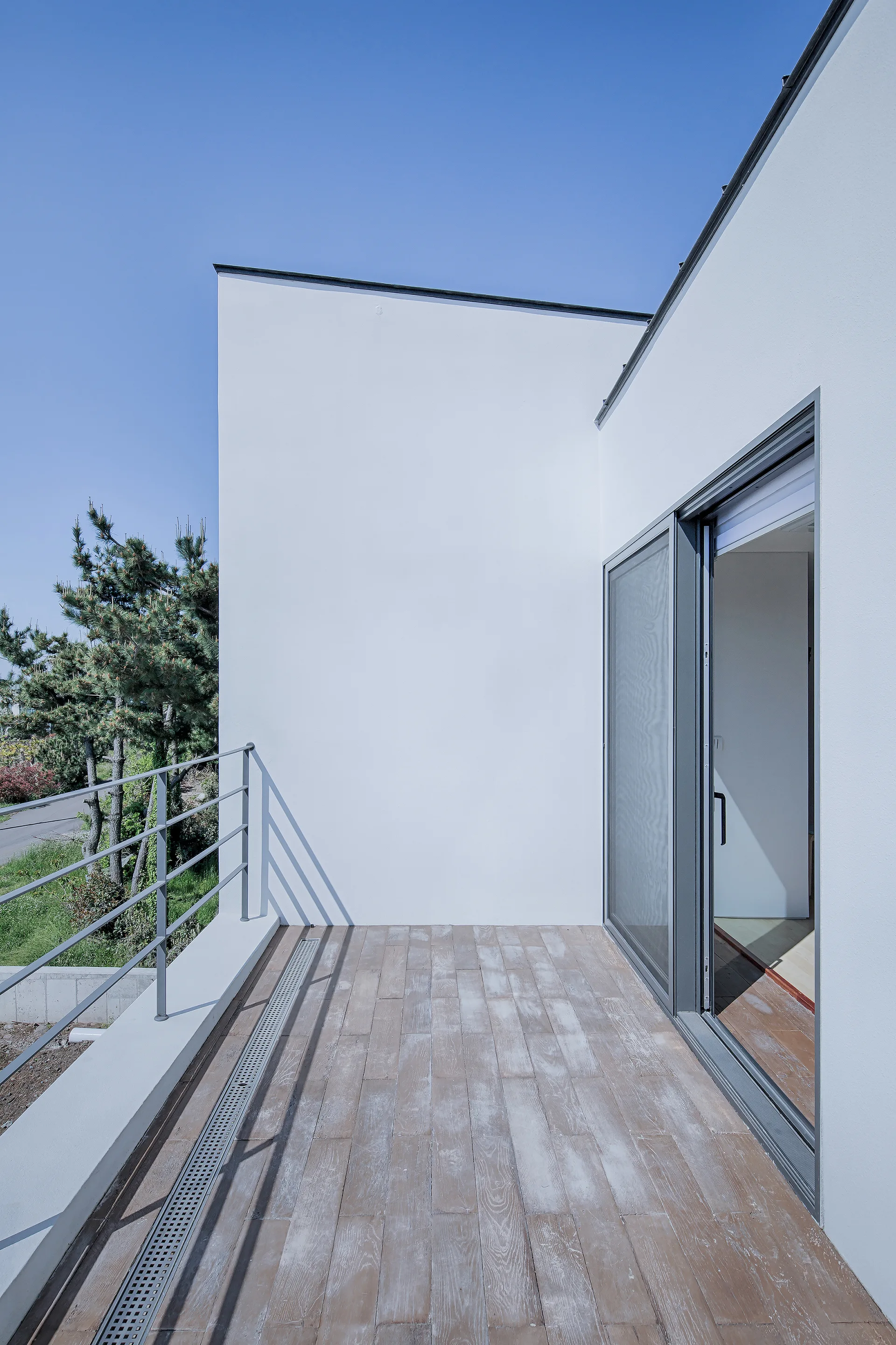 A modern minimalist balcony features weathered wood-look plank flooring, white stucco walls, a grey metal railing, and a sliding glass door.