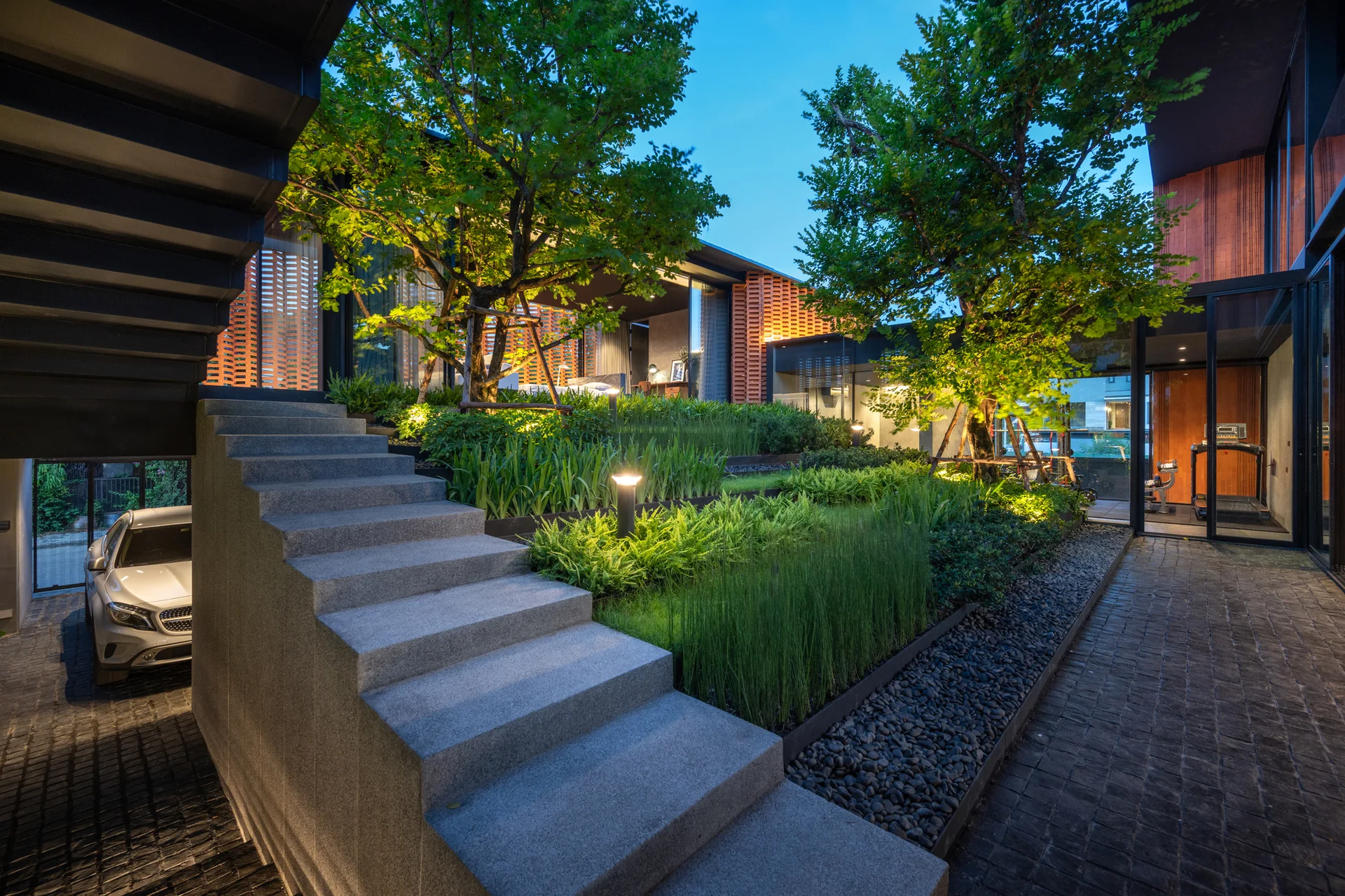 A modern courtyard features a multi-tiered terraced garden illuminated by bollard lights, adjacent to a solid stone staircase and dark cobblestone path.