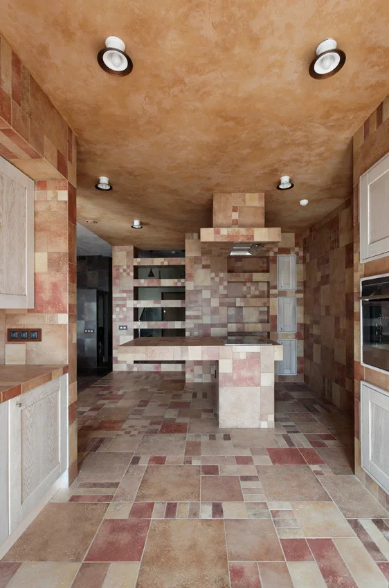 A modern kitchen featuring earth-toned patchwork tiles covering the walls, floor, and central island beneath a warm plaster ceiling.