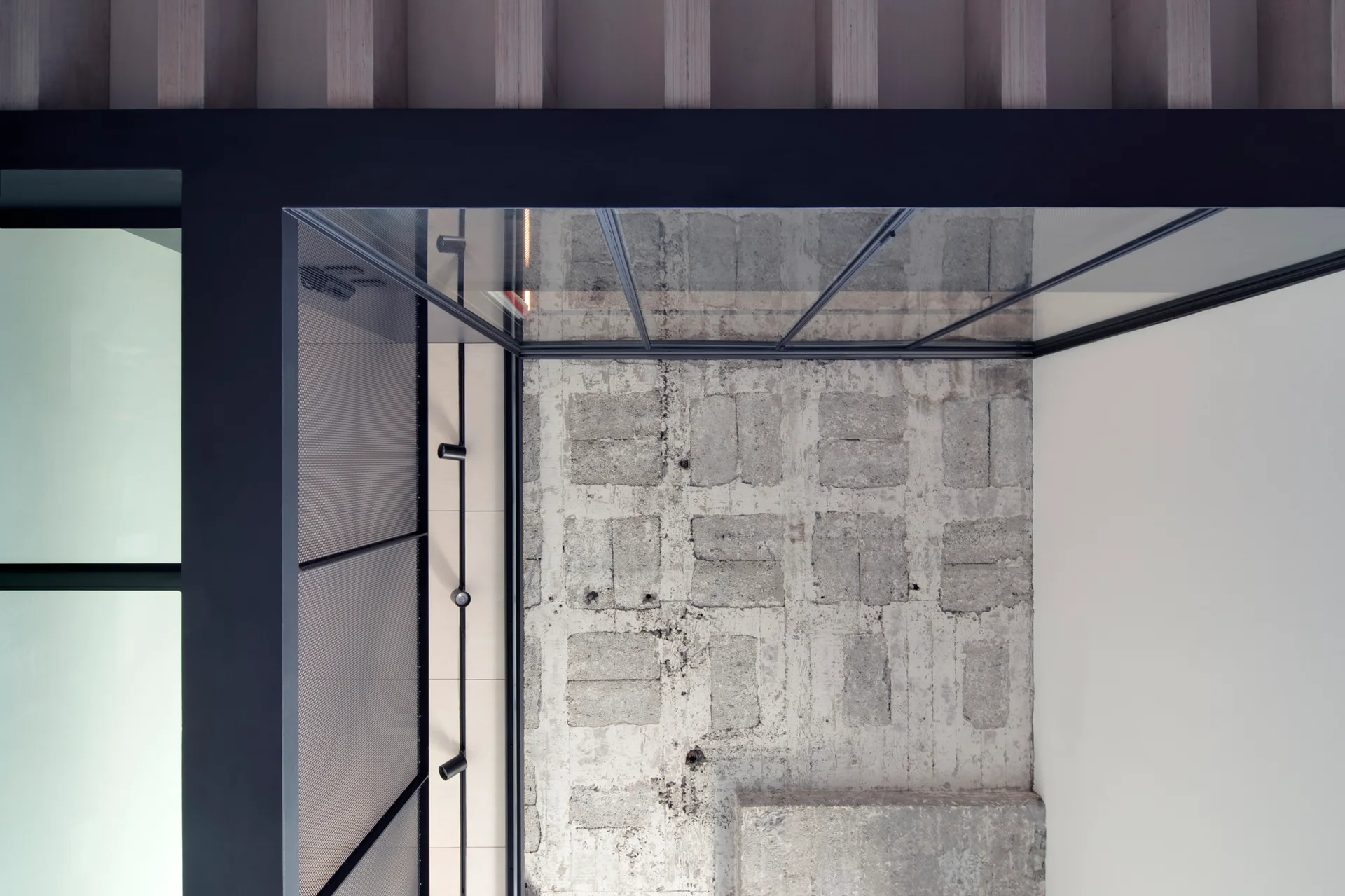 Architectural detail of an interior ceiling featuring a glass panel frame, exposed concrete block wall, black track lighting, and vertical wood slats.