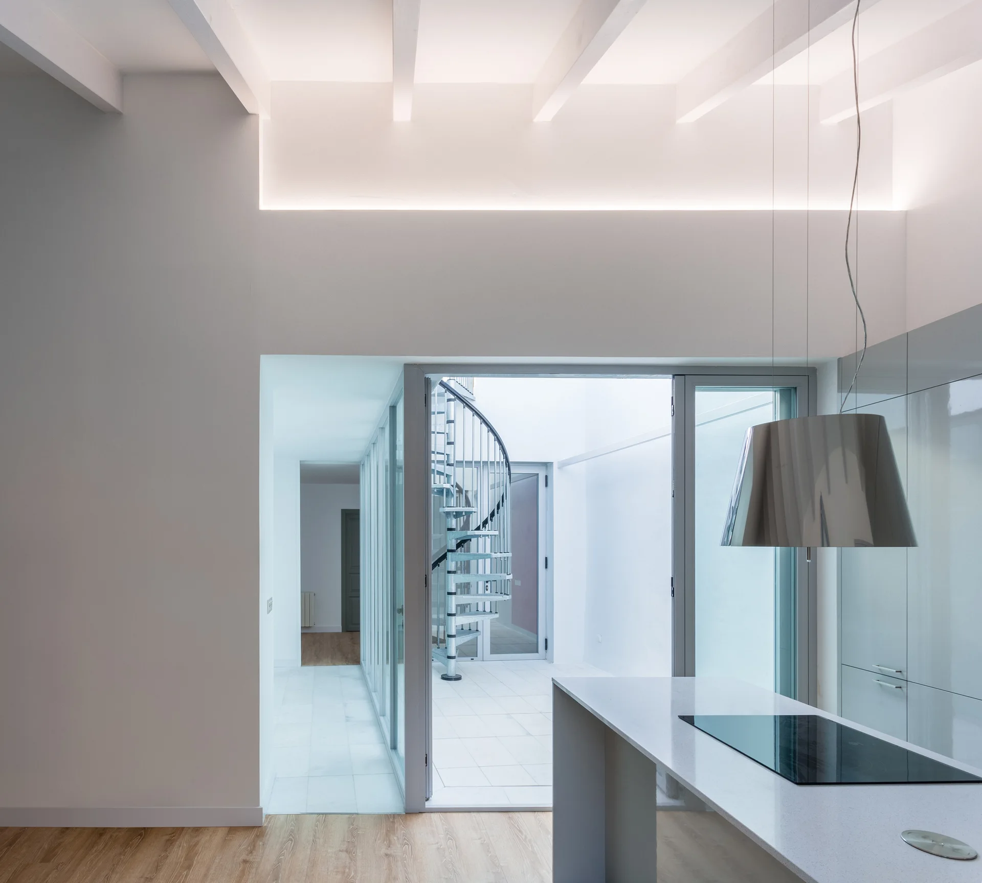A modern white kitchen features a sleek island with a cooktop, looking out through glass doors to a metal spiral staircase in a bright courtyard.