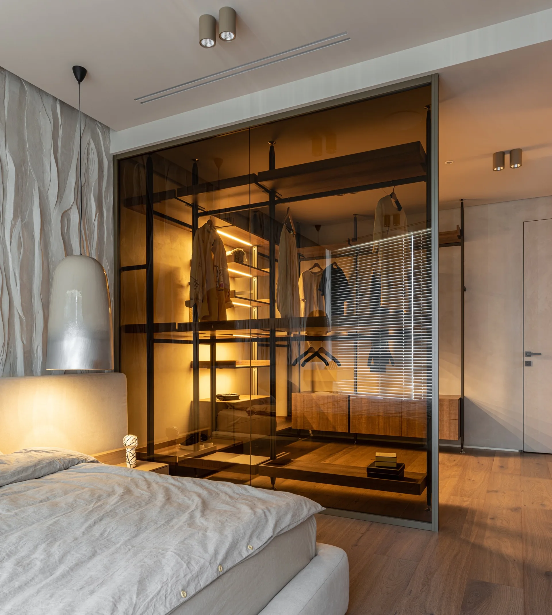 A modern bedroom features an upholstered bed beside a textured wall and a walk-in wardrobe enclosed by tinted glass panels with illuminated shelves.