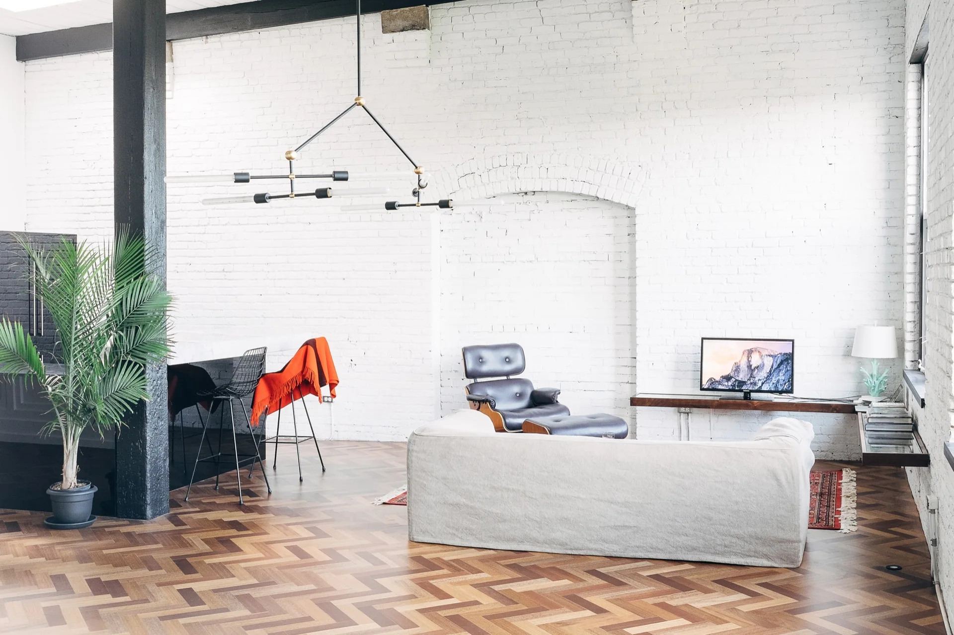 A bright industrial living room features white painted brick walls, herringbone wood flooring, a cream sofa, and a classic black leather lounge chair.