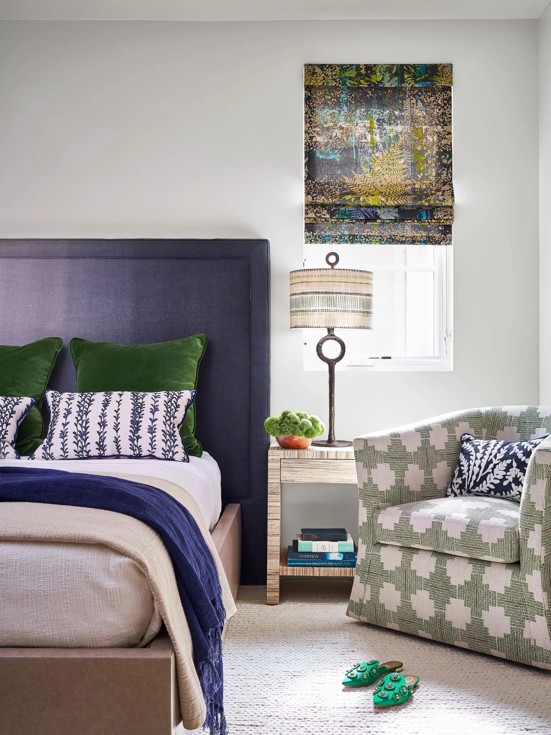 A bedroom corner features a dark navy headboard, a patterned cream-and-green armchair, a textured side table, and a patterned Roman window shade.