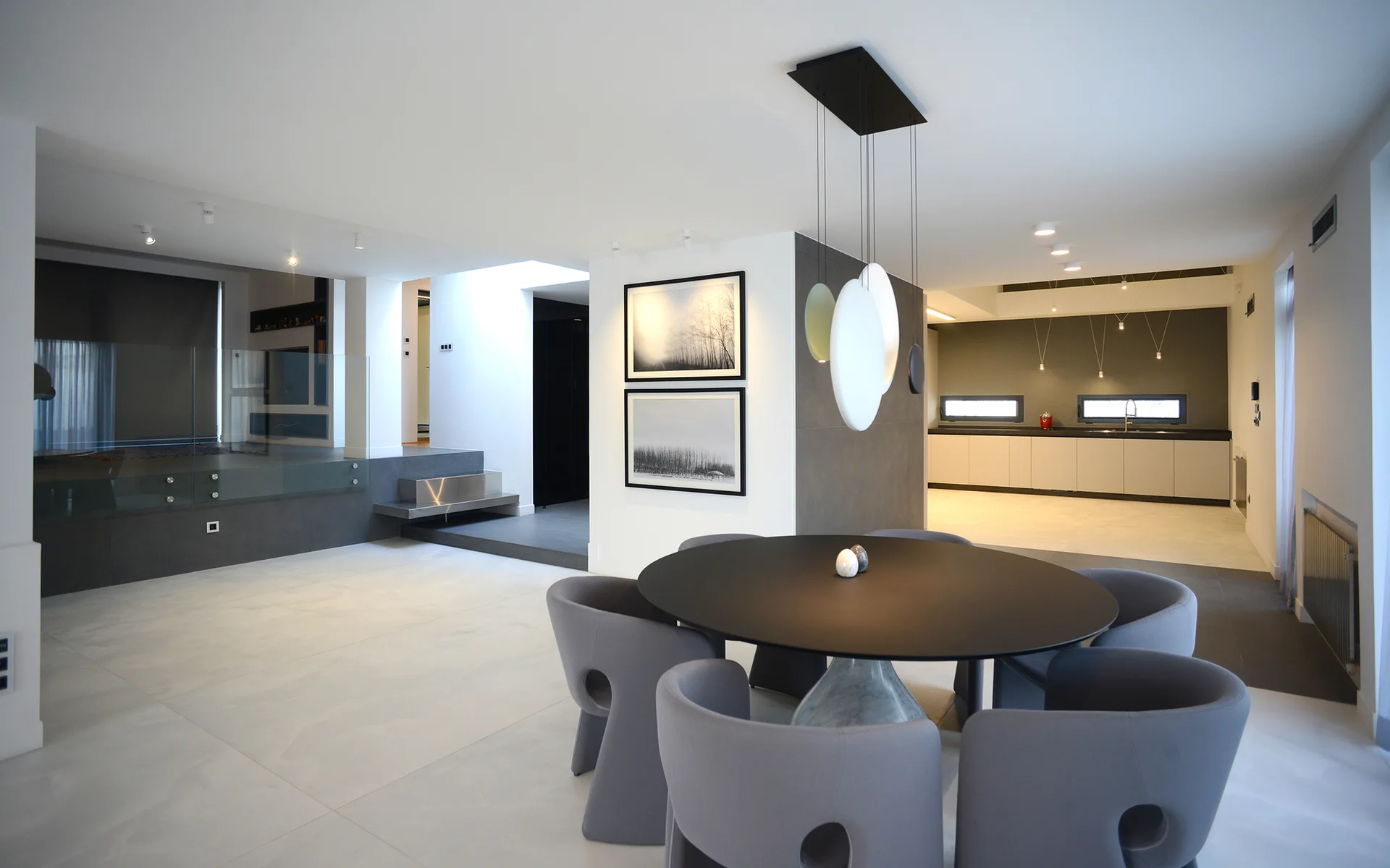 A minimalist dining room features a round black table surrounded by grey cutout chairs, under hanging disc pendant lights in an open-plan home.
