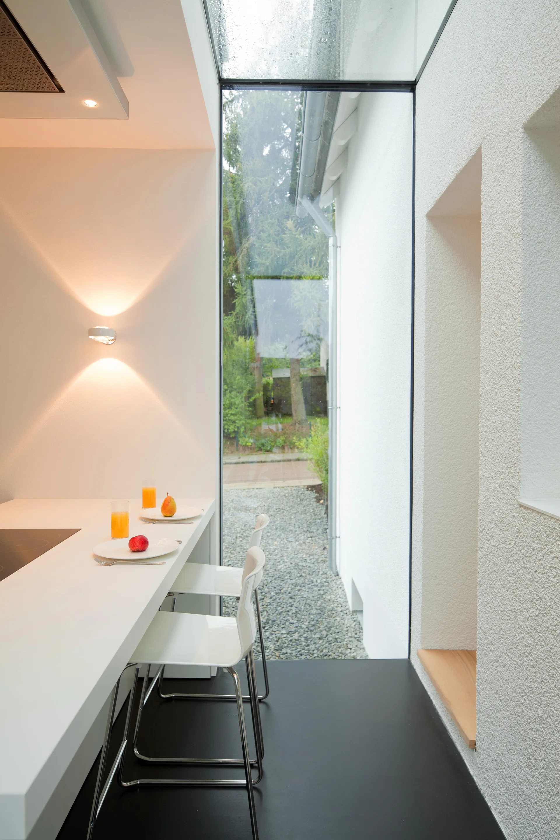 A minimalist kitchen features a white counter with bar stools, a black floor, and a tall glass wall opening up to a gravel path outside.