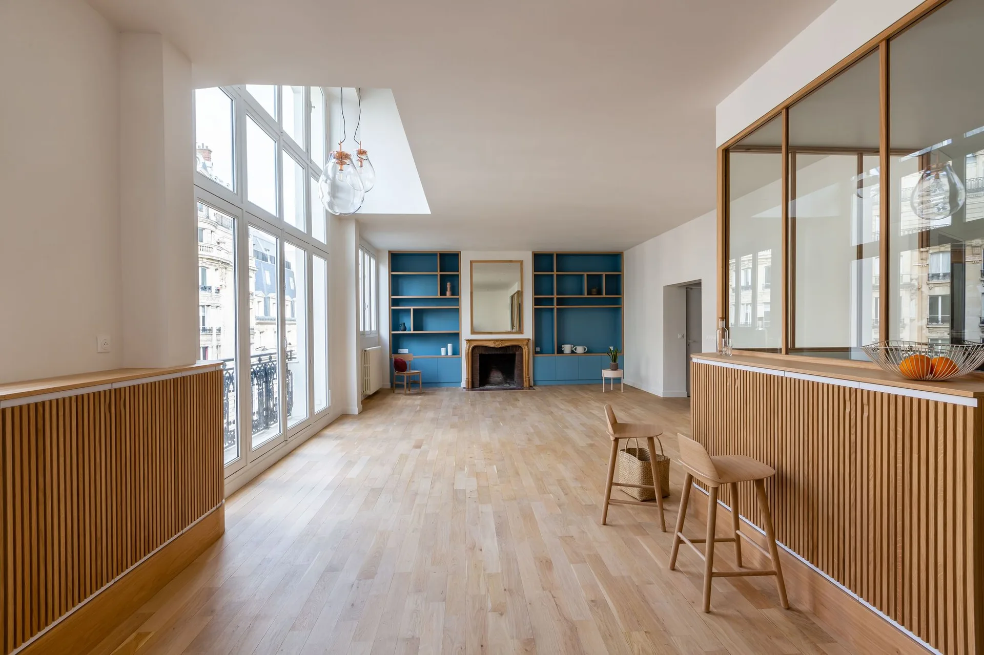 A spacious room features light oak floors, a blue built-in shelving unit surrounding a fireplace, and vertical wood slat paneling.