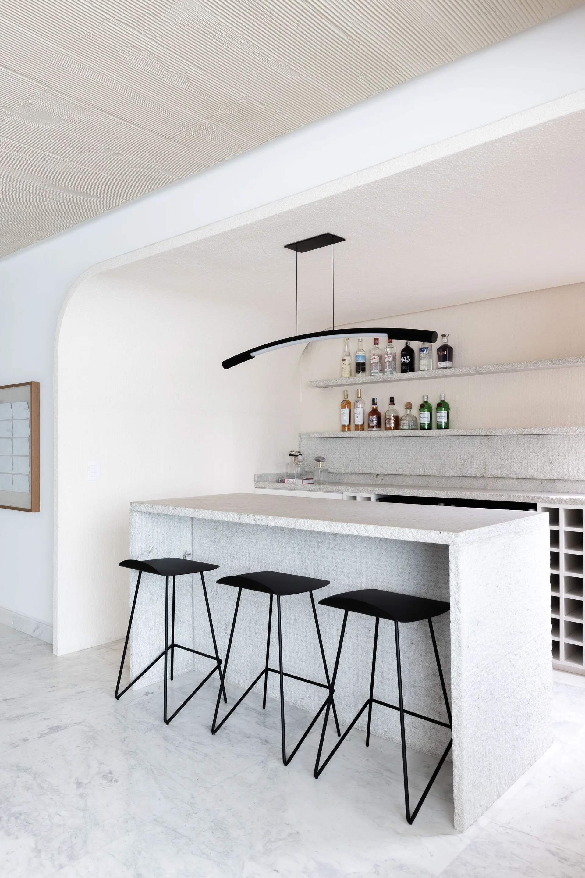 A minimalist home bar features a rough stone slab counter with three black stools beneath a curved alcove and a black pendant light.