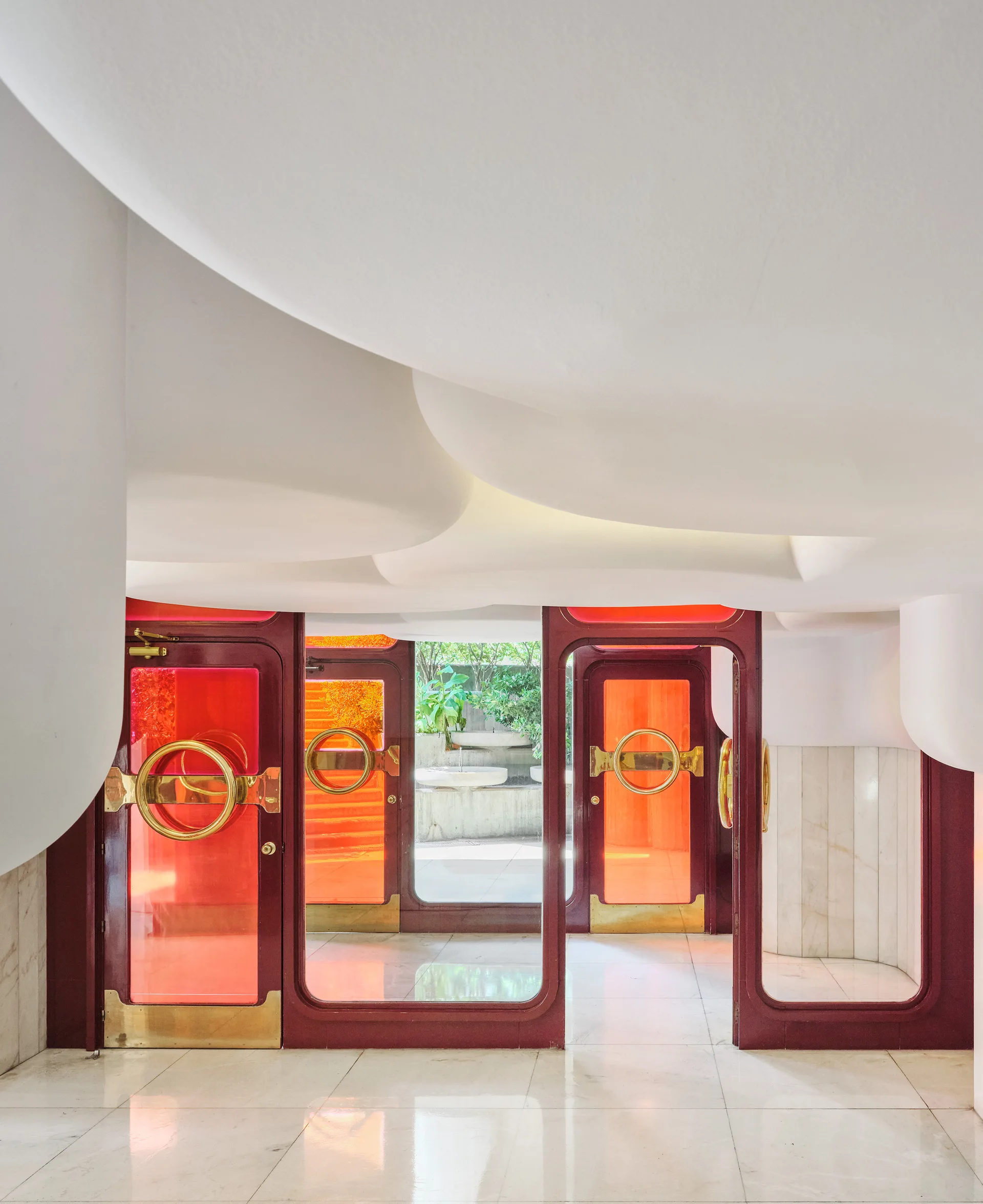 An entryway features curved, organic white ceiling forms above red-tinted glass doors with circular gold handles and polished floor tiles.