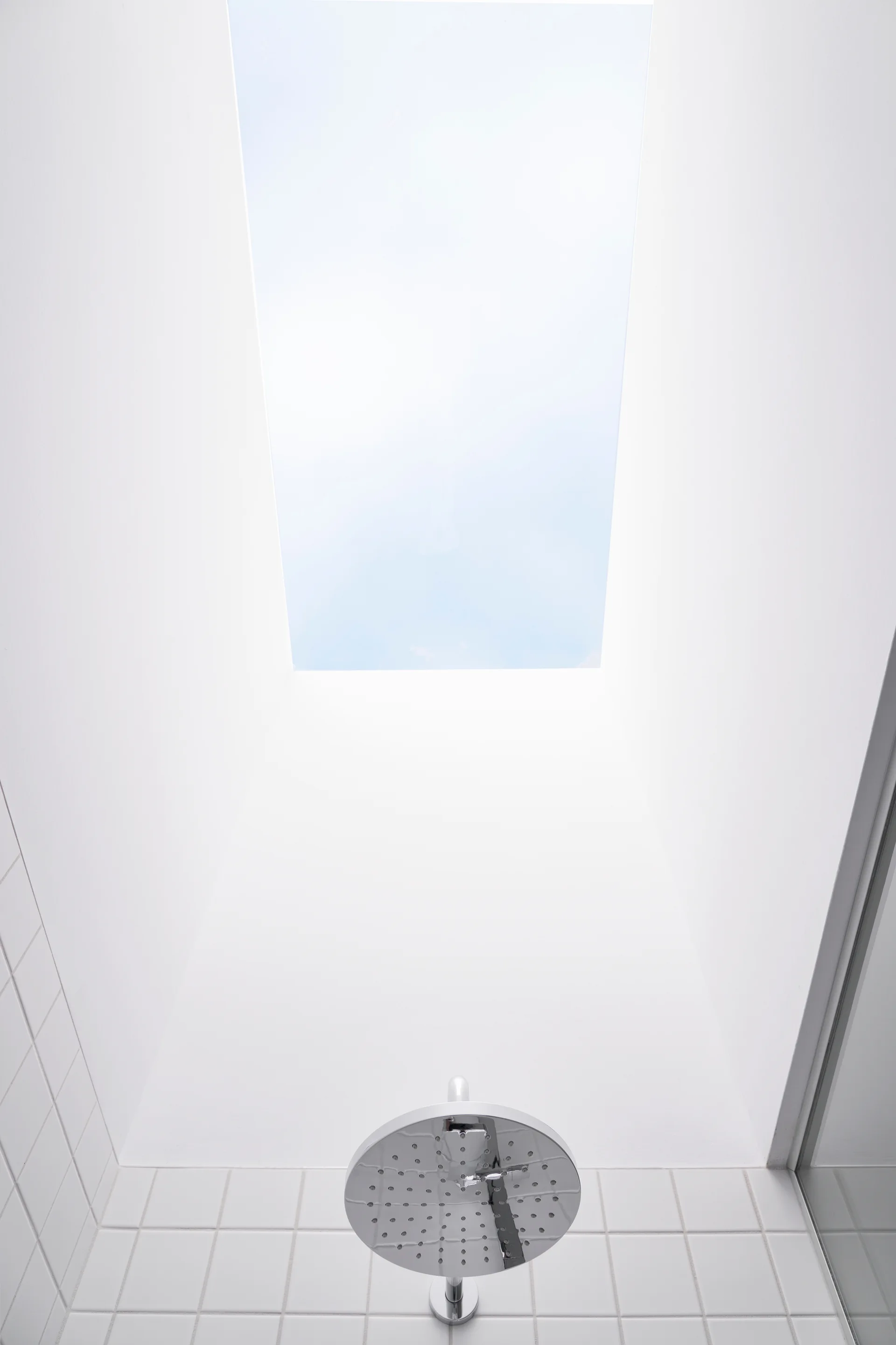 A low-angle view of a circular chrome shower head mounted on a white tiled wall beneath a large rectangular skylight showing the sky.