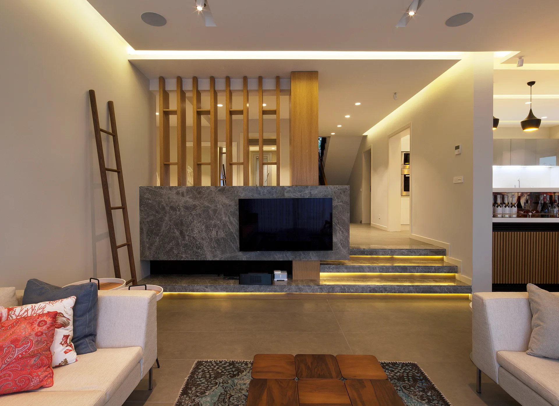 A contemporary living room shows a gray marble media wall and lighted stairs next to a vertical wood slatted screen and cream sofas.