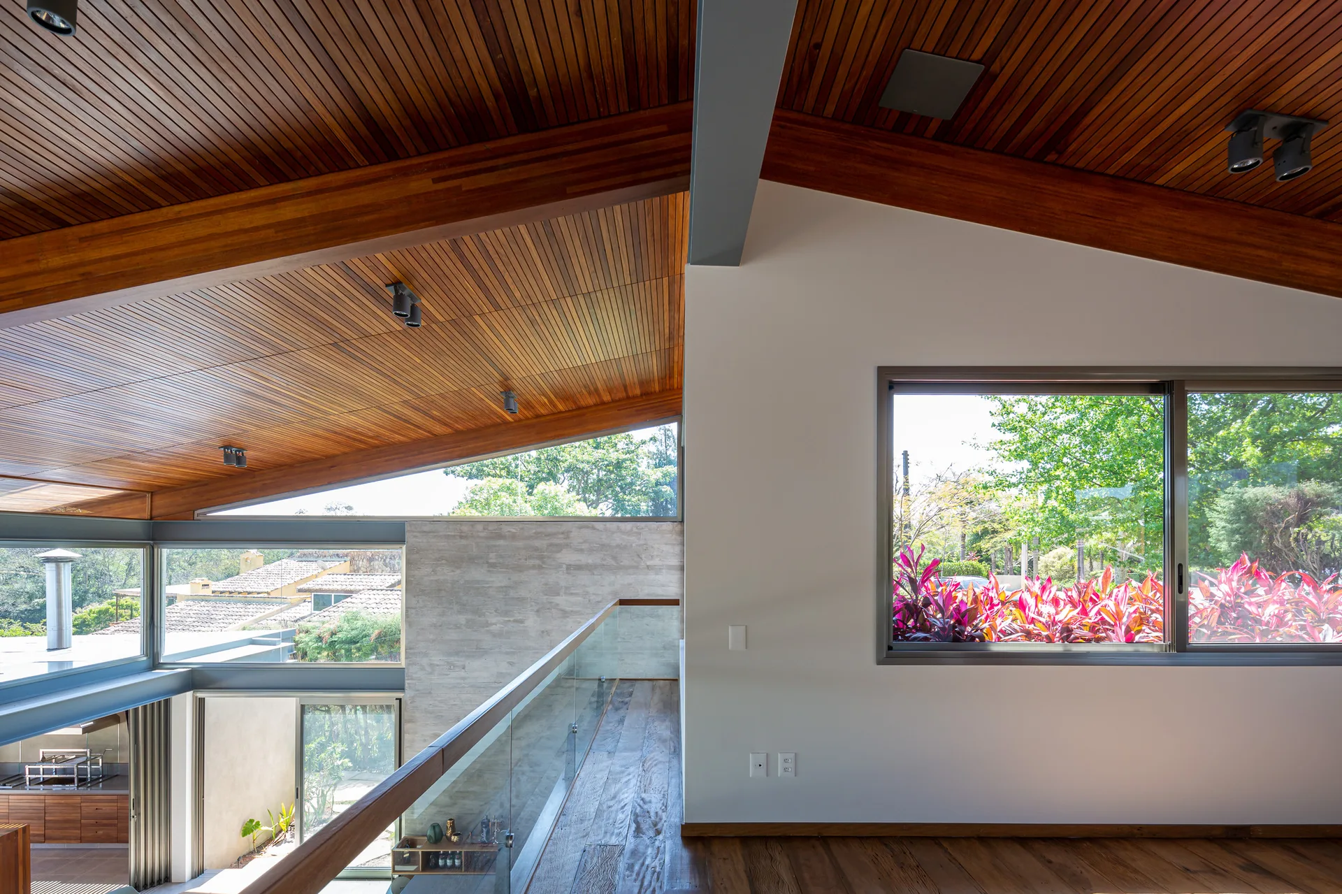 A modern mezzanine landing features a vaulted wood-paneled ceiling, a glass panel railing, and large windows looking out to trees.