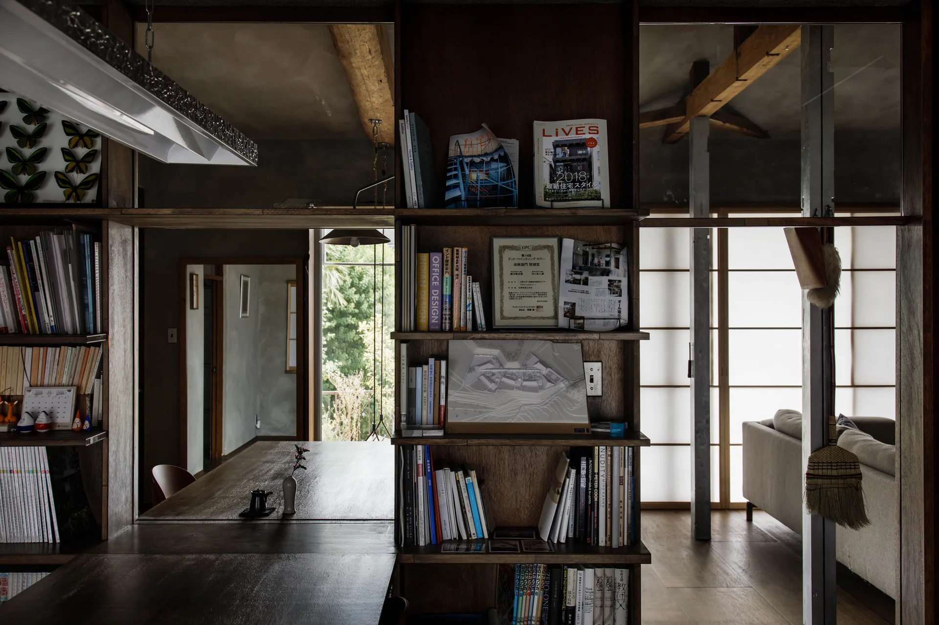 A dark wood bookshelf partition divides a room featuring exposed ceiling beams, a dining table, and translucent shoji screens.