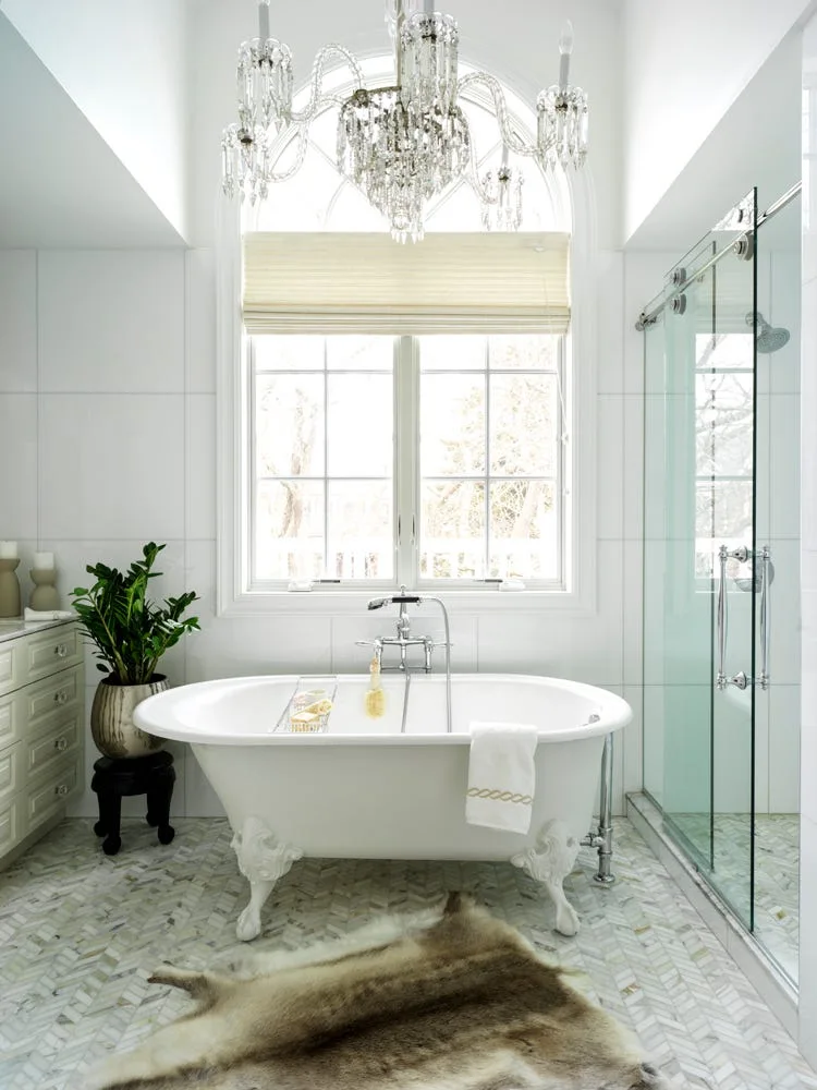 A white clawfoot bathtub sits on herringbone marble tile beneath a crystal chandelier and an arched window in a bright bathroom.