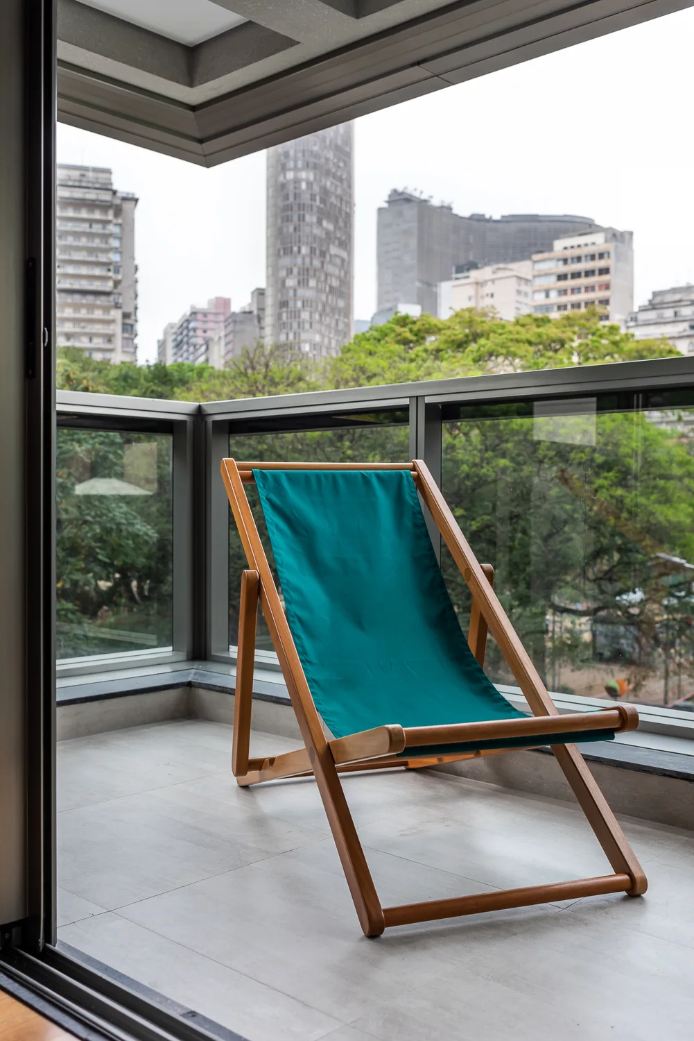 A wooden folding deck chair with a teal canvas seat sits on a tiled balcony with a glass railing overlooking a city skyline.