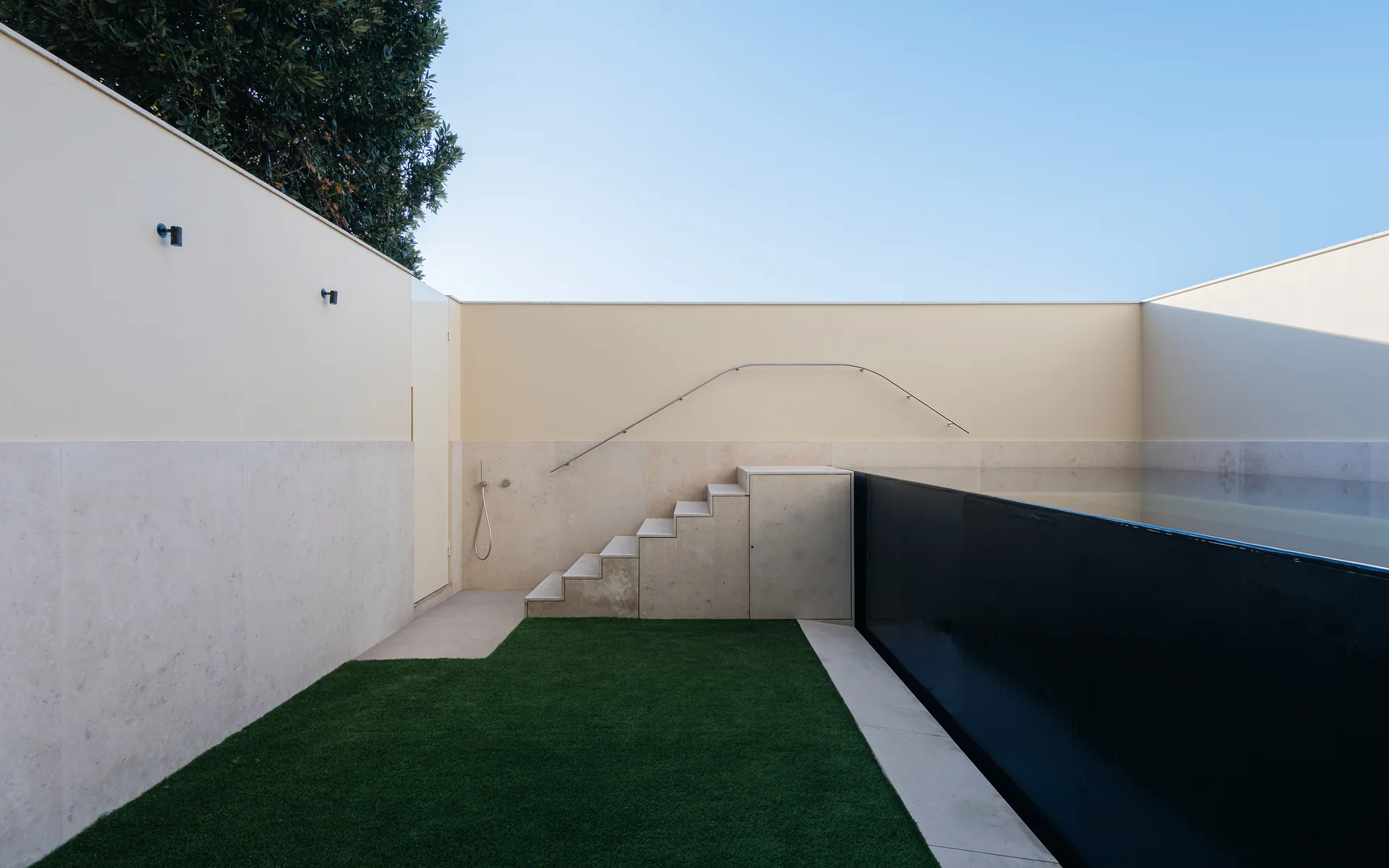 A minimalist outdoor terrace features an elevated black-walled pool, beige stone steps, a synthetic green turf lawn, and high cream-colored perimeter walls.