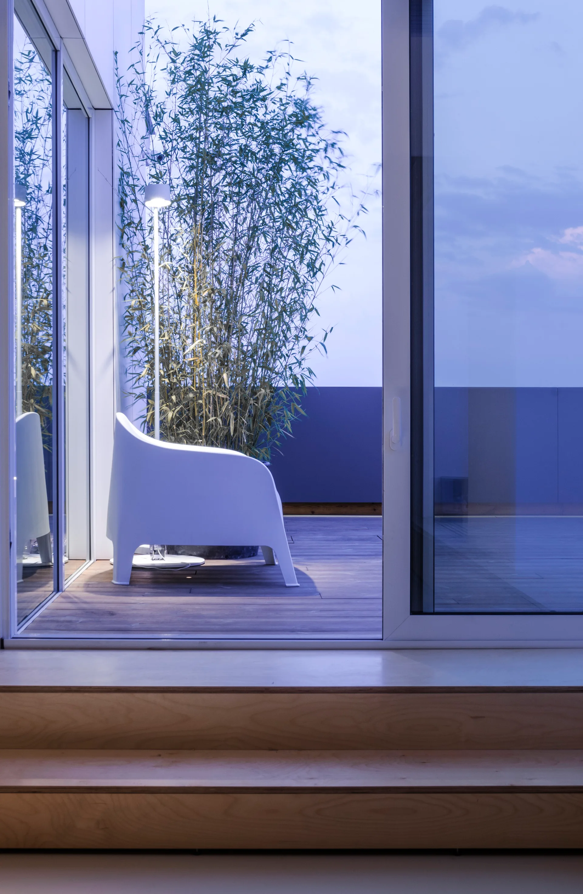A white modern armchair sits beside tall potted bamboo and a slender floor lamp on a wooden balcony deck seen through a sliding glass door.