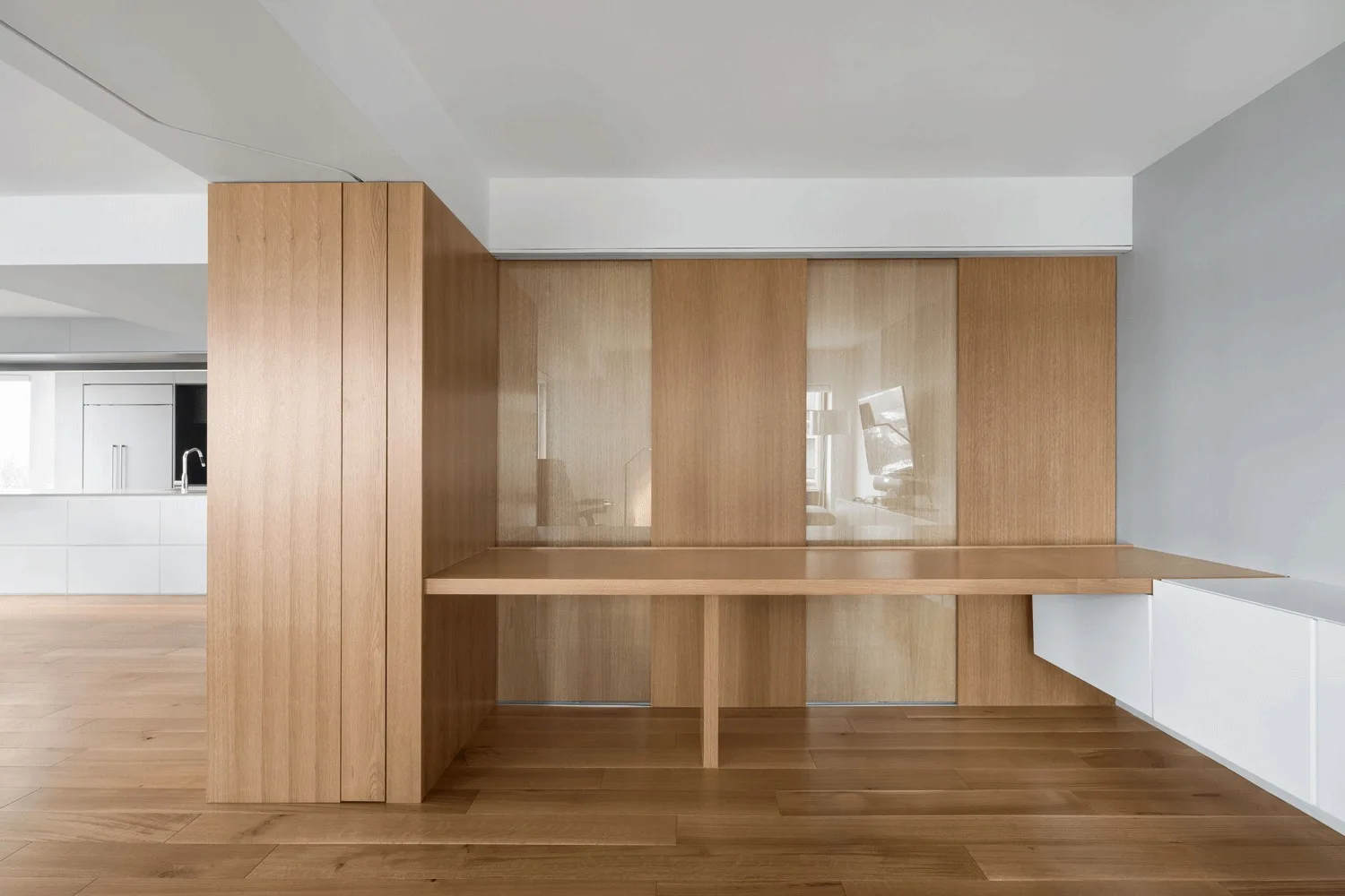 A minimalist home office area features a long built-in wooden desk integrated with floor-to-ceiling timber cabinetry and sliding panels.