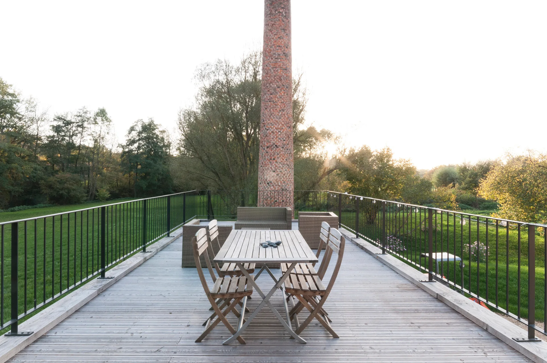 An outdoor wooden deck with a central wood dining table, surrounding folding chairs, wicker lounge sofas, and a tall brick chimney in the background.