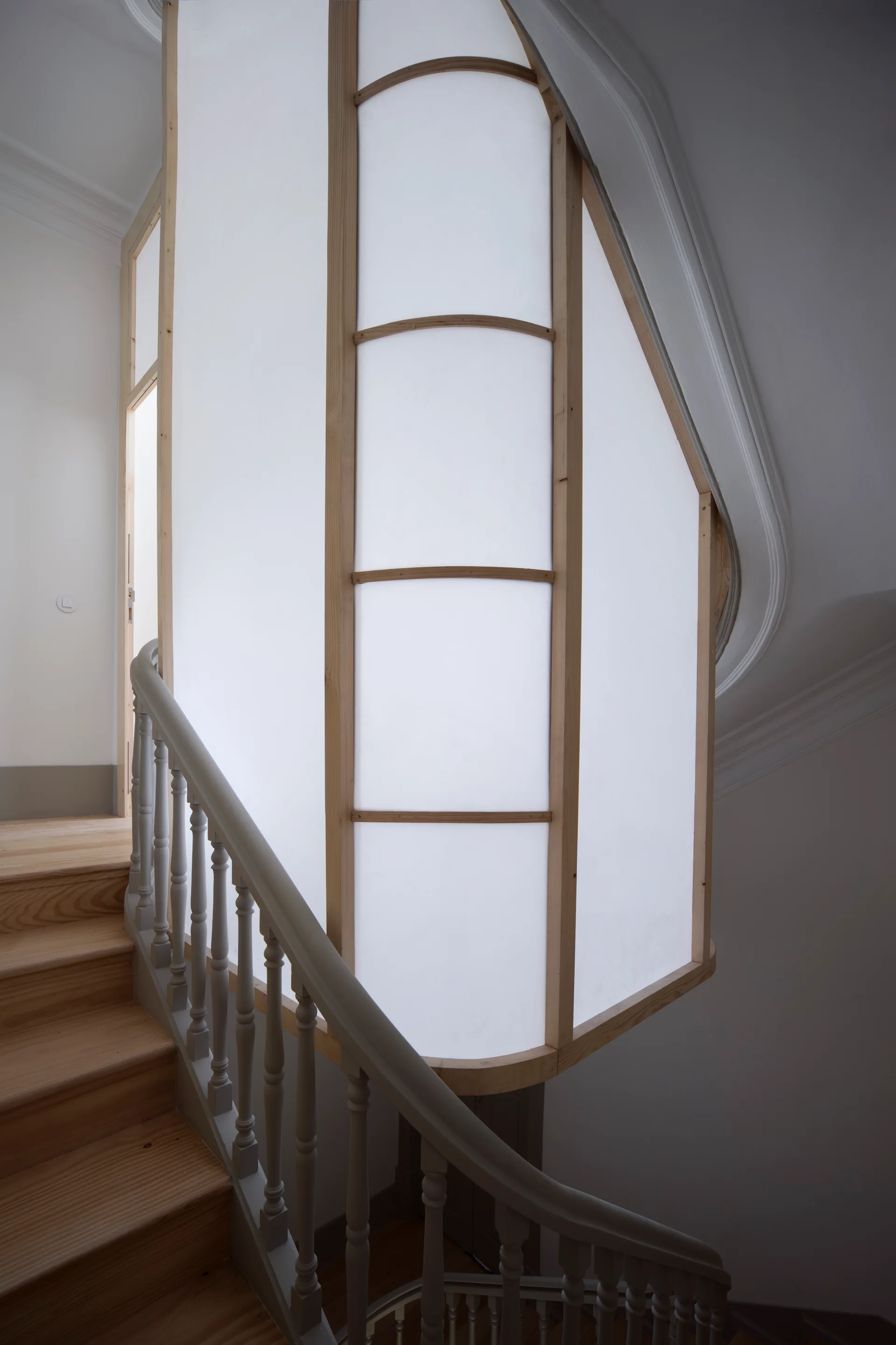 A curved wooden staircase with grey balusters stands beside a large, illuminated translucent panel framed in light wood.