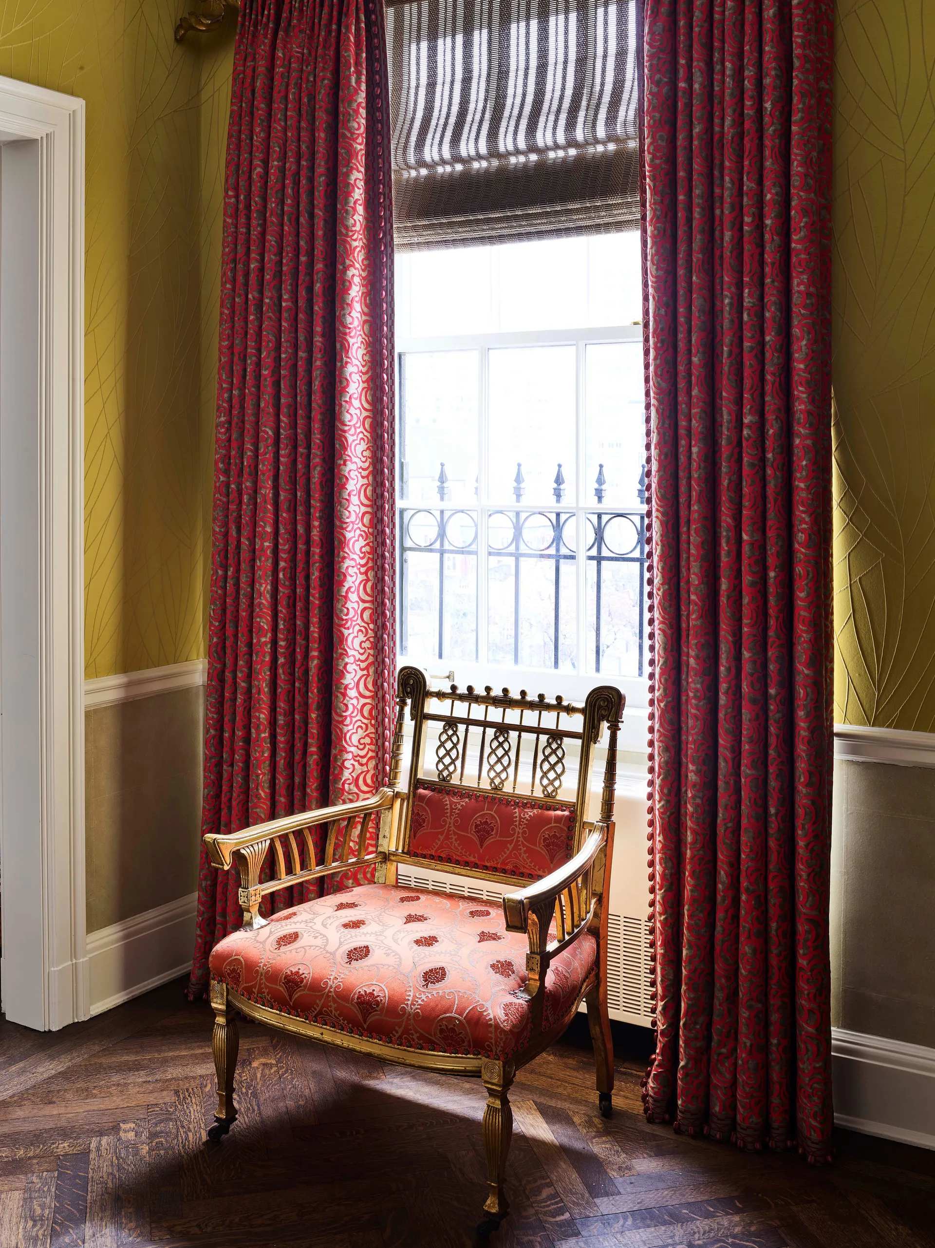 An ornate gold armchair sits in a room corner beside red patterned drapes, a large window with a striped shade, and olive-green wallpaper.