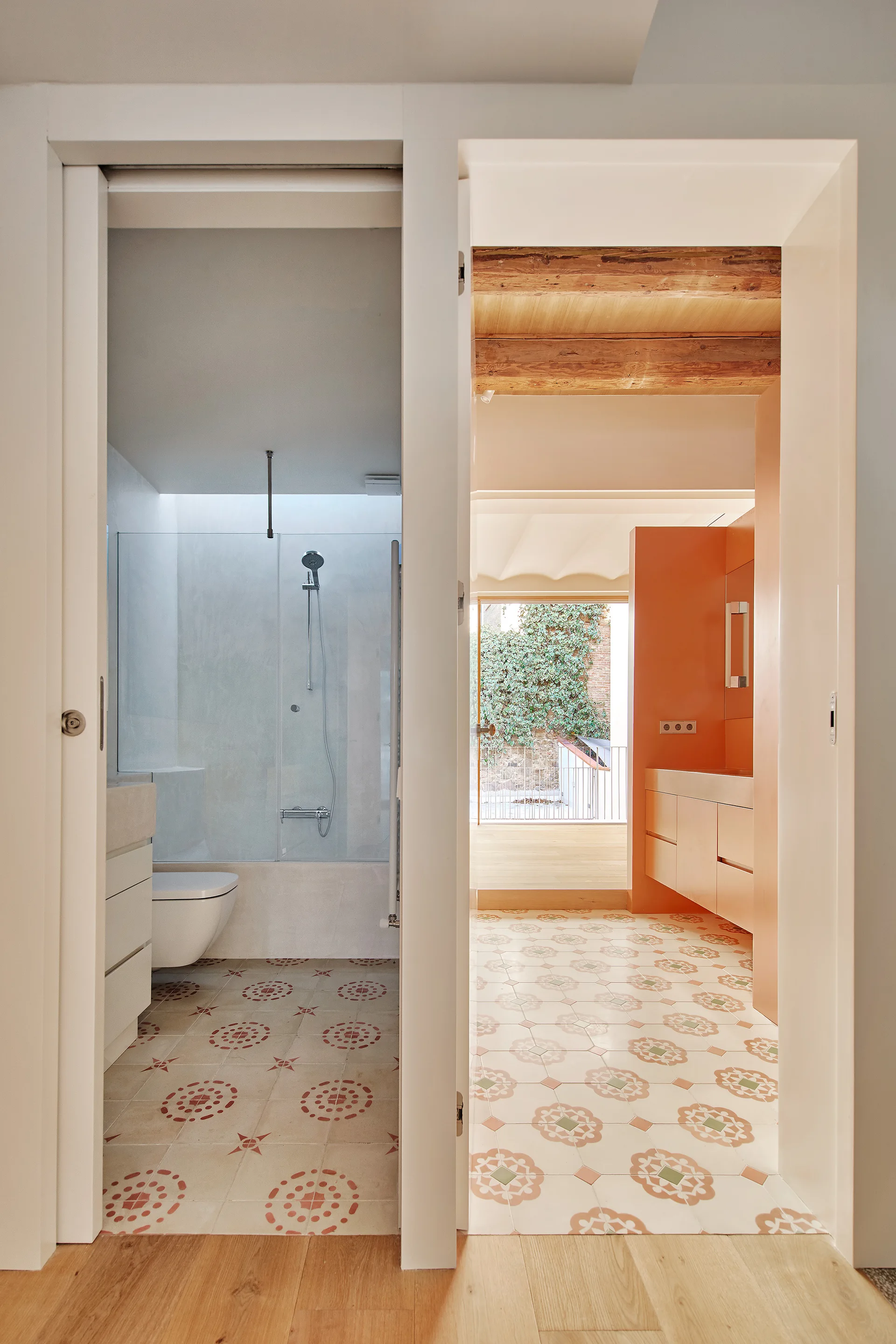 A split-view doorway reveals a bathroom with a glass shower and a vanity area featuring orange cabinetry, wood ceiling beams, and patterned floor tiles.