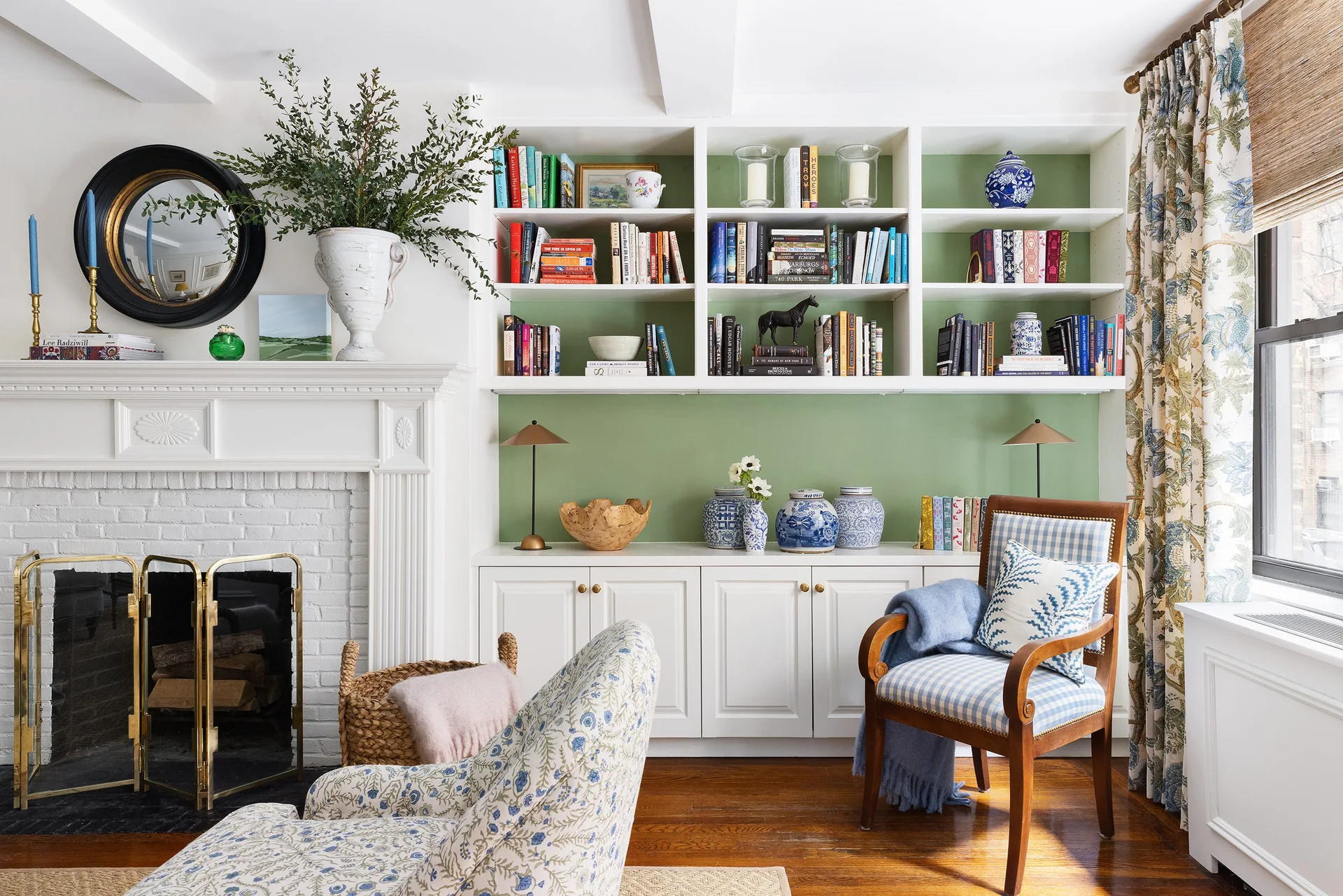 A traditional living room features a white brick fireplace, built-in white bookshelves with green backing, and a blue and white checkered armchair.