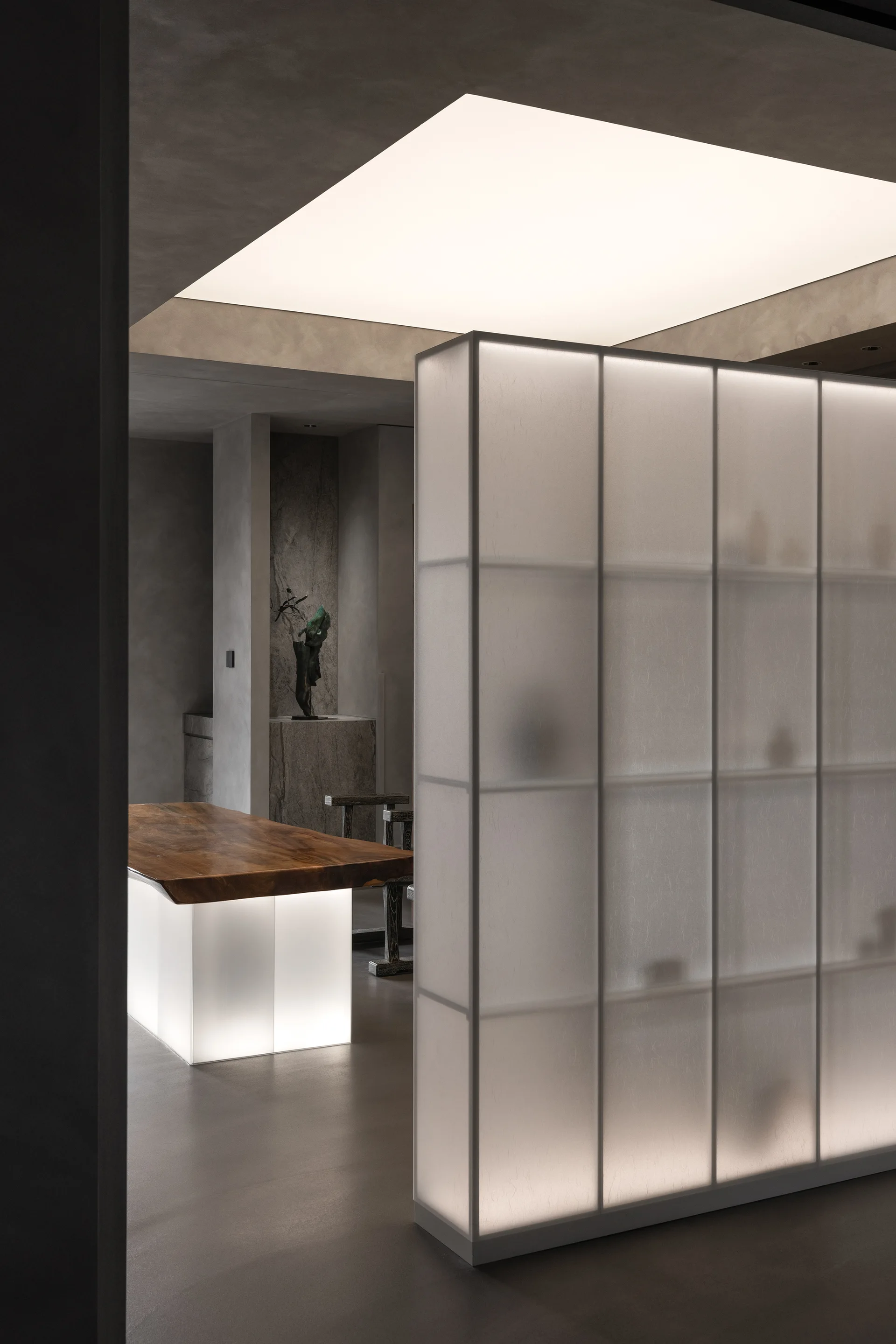 A minimalist gray room features a tall backlit frosted glass partition and a live-edge wood table with a glowing base.