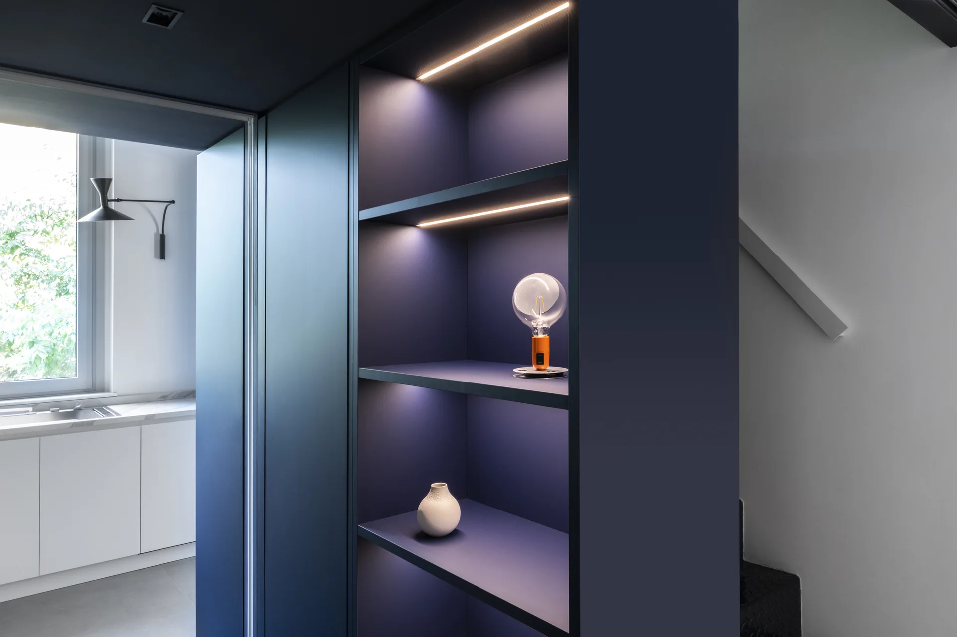 A matte dark blue built-in shelving unit features integrated LED strip lights, a white ceramic vase, and a sculptural orange lamp.