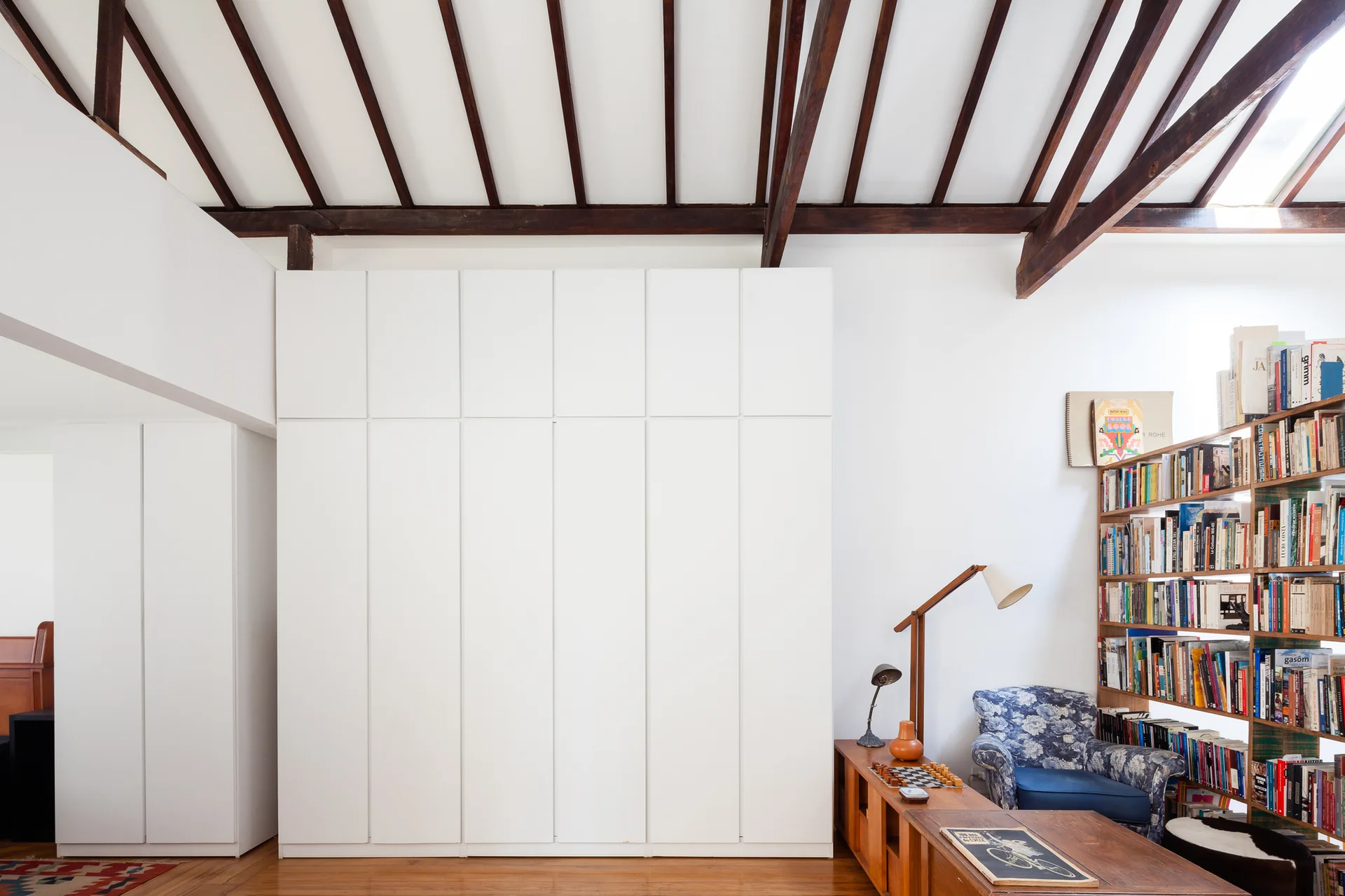 A living room features a large floor-to-ceiling white wardrobe beneath dark exposed wooden ceiling beams next to a wooden bookcase and armchair.