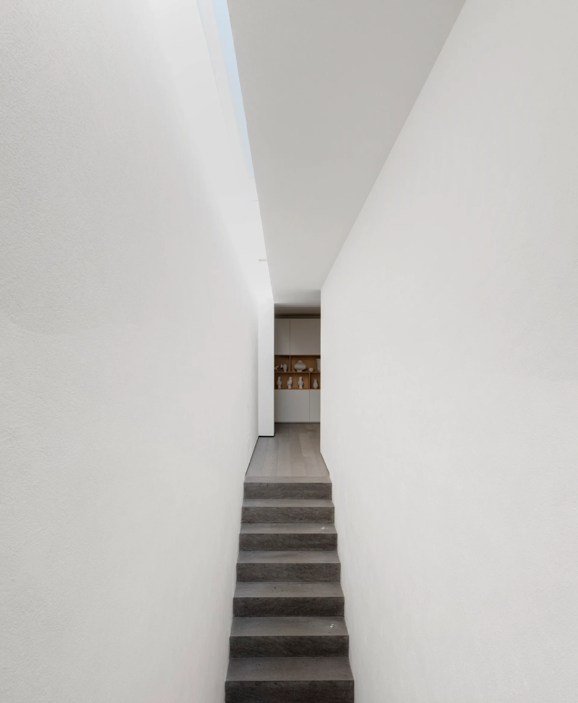 A narrow white minimalist corridor features dark grey stone stairs leading to a wood-floored landing with recessed display shelving.
